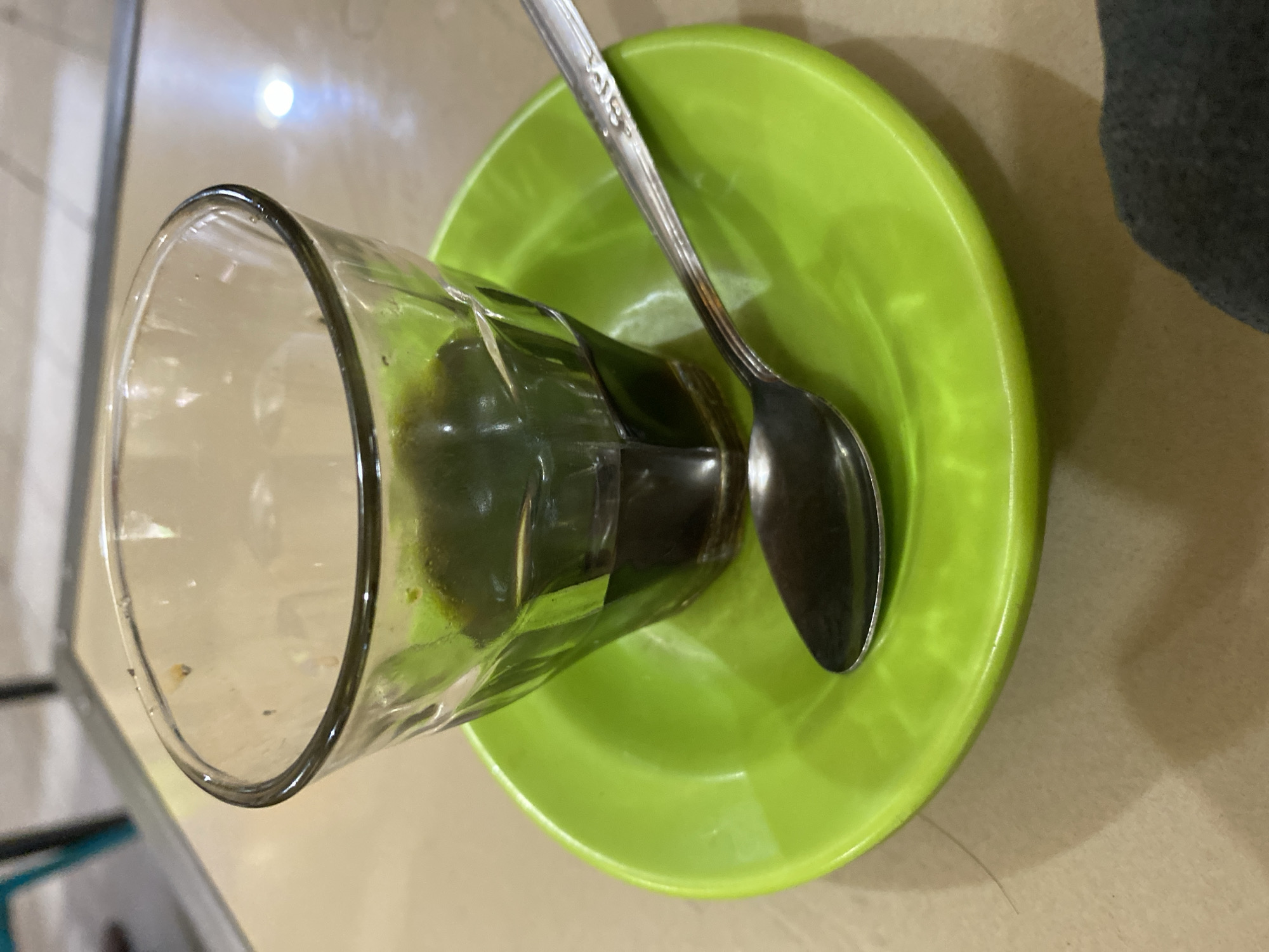 A close-up, eye-level shot shows a nearly empty glass of dark liquid, likely coffee or tea, sitting on a bright green saucer. A silver spoon rests on the saucer, angled towards the lower right of the frame. The glass appears to have small dark specks on its rim. The saucer is slightly reflective, showing highlights from overhead lighting. The background consists of a light-colored, textured surface, possibly a table, with a blurred background that hints at furniture or structural elements. The scene is devoid of people and specific location indicators, making it impossible to ascertain its presence in Lhokseumawe, Indonesia, or any other city.