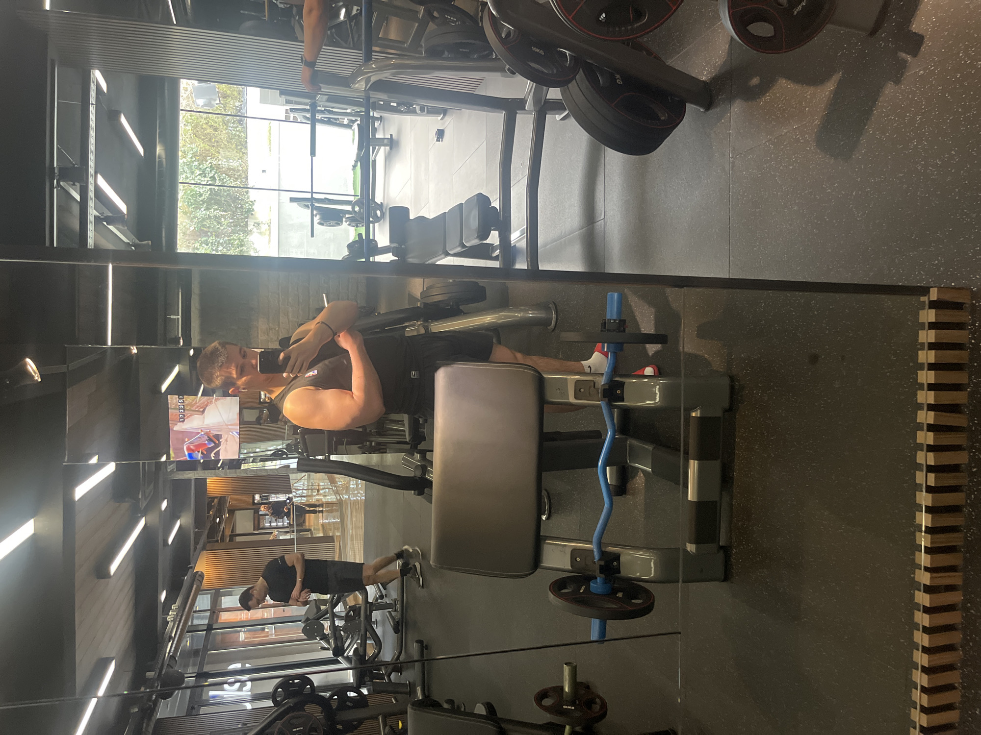 This image depicts the interior of a modern fitness facility, specifically a gym, captured via a self-portrait taken in a large, reflective wall mirror.
**People:**
The primary subject is a male, estimated to be in his twenties to thirties, positioned centrally and slightly to the right of the frame. He is taking a selfie with a dark-colored smartphone held in his left hand, while flexing his right bicep. He wears a dark sleeveless top and dark athletic shorts. His footwear consists of white socks paired with red slide sandals.
In the reflected background, to the left, a second male, estimated to be in his thirties to forties, is visible. He wears a black t-shirt, dark shorts, glasses, and white earbuds, and is looking down at his left wrist, possibly checking a smartwatch. He appears to be walking from left to right.
A third, less distinct individual is partially visible further in the background, near an entrance or reception area.
**Subjects/Objects:**
In the immediate foreground, a grey preacher curl bench with a blue EZ bar is prominent. The EZ bar is loaded with black weight plates, at least one of which appears to be a 10kg plate. Various other gym equipment, including weight machines, racks, and benches, are reflected throughout the gym space. A large flat-screen display, reflected in the upper right background, shows digital content, possibly a video game or sports feed, with numerical data "17 23 59" visible.
**Actions/Interactions:**
The primary action is the self-portrait being taken by the man in the foreground. The second man is checking his wrist. No direct interactions between individuals are depicted.
**Environment:**
The gym features a dark, speckled rubber floor. The ceiling is dark, equipped with multiple rectangular LED strip lights. The architectural design incorporates sections of dark, exposed brick-like wall texture and vertical wooden slat paneling. Large windows along one wall reveal an outdoor view of lush green foliage, suggesting an adjacent garden or natural area. A faint white logo, "Pulse," is partially discernible on a reflected glass surface in the mid-background, indicating the facility's branding.
**Location in Sofia, Bulgaria:**
The branding "Pulse" identifies this as a **Pulse Fitness & Spa** gym. Based on the distinctive modern architectural elements, including the wooden slat paneling, spacious layout, and large windows overlooking greenery, the specific location is identified as **Pulse Fitness & Spa Mladost** in **Sofia, Bulgaria**.