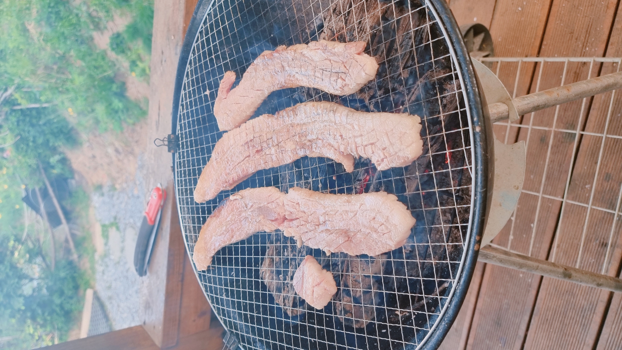 The image shows a grill with pieces of meat cooking on it. The meat is pale and appears to be pork belly or a similar fatty cut. The grill is a standard round charcoal grill with a wire grate. Smoke is rising from the grill, indicating that the charcoal is burning. A grill tool sits on the side of the grill. The grill is situated on a wooden deck or patio. In the background, green foliage suggests a yard or garden.
The location is Dunnae-myeon, South Korea.