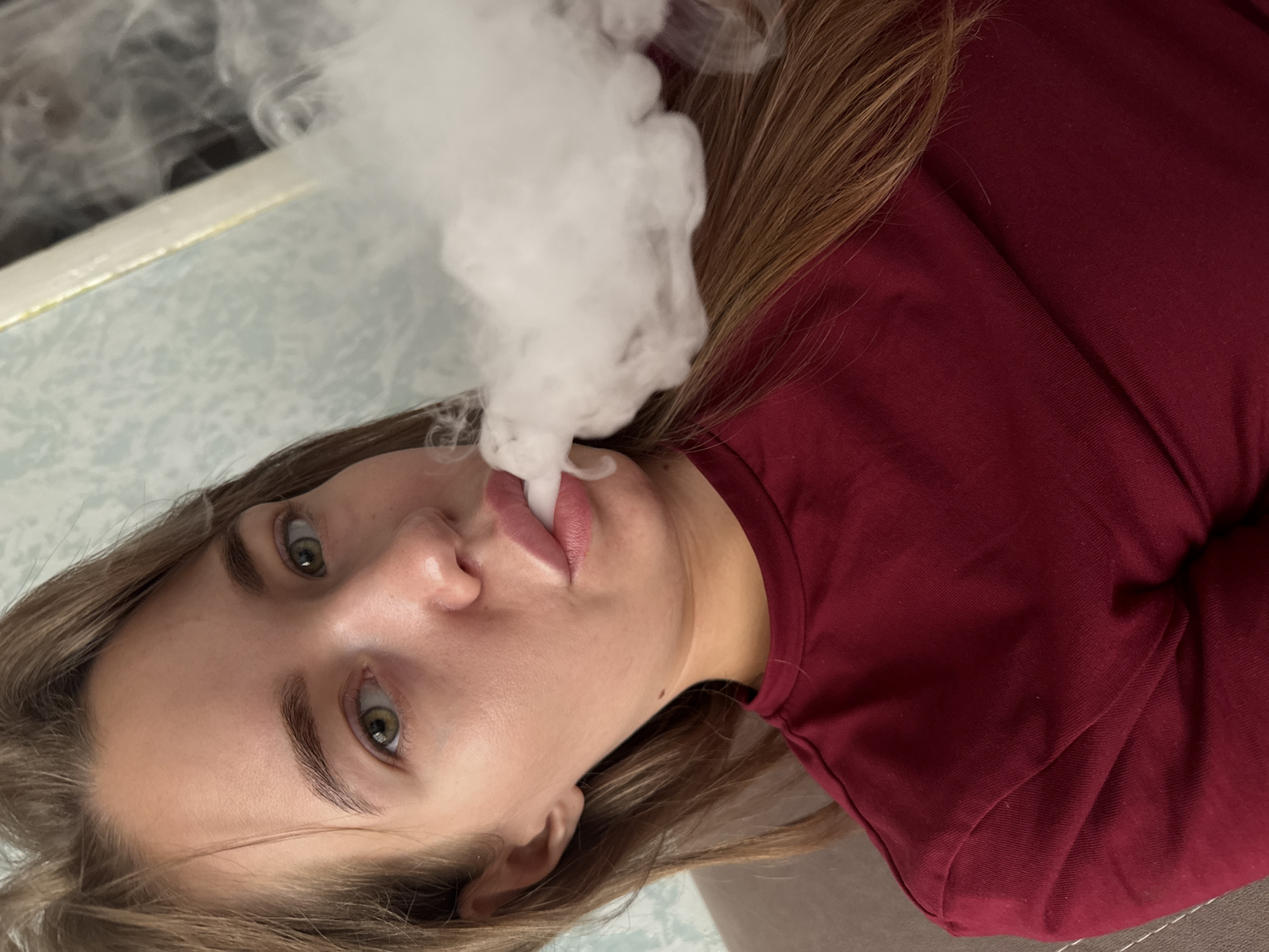 The image shows a young woman with long brown hair and green eyes, wearing a maroon t-shirt. She is exhaling a cloud of vapor. The background consists of a patterned wallpaper and a sofa. The image is a close-up selfie, and no specific location in the city of Chaikovsky, Russia can be determined from the visual information.