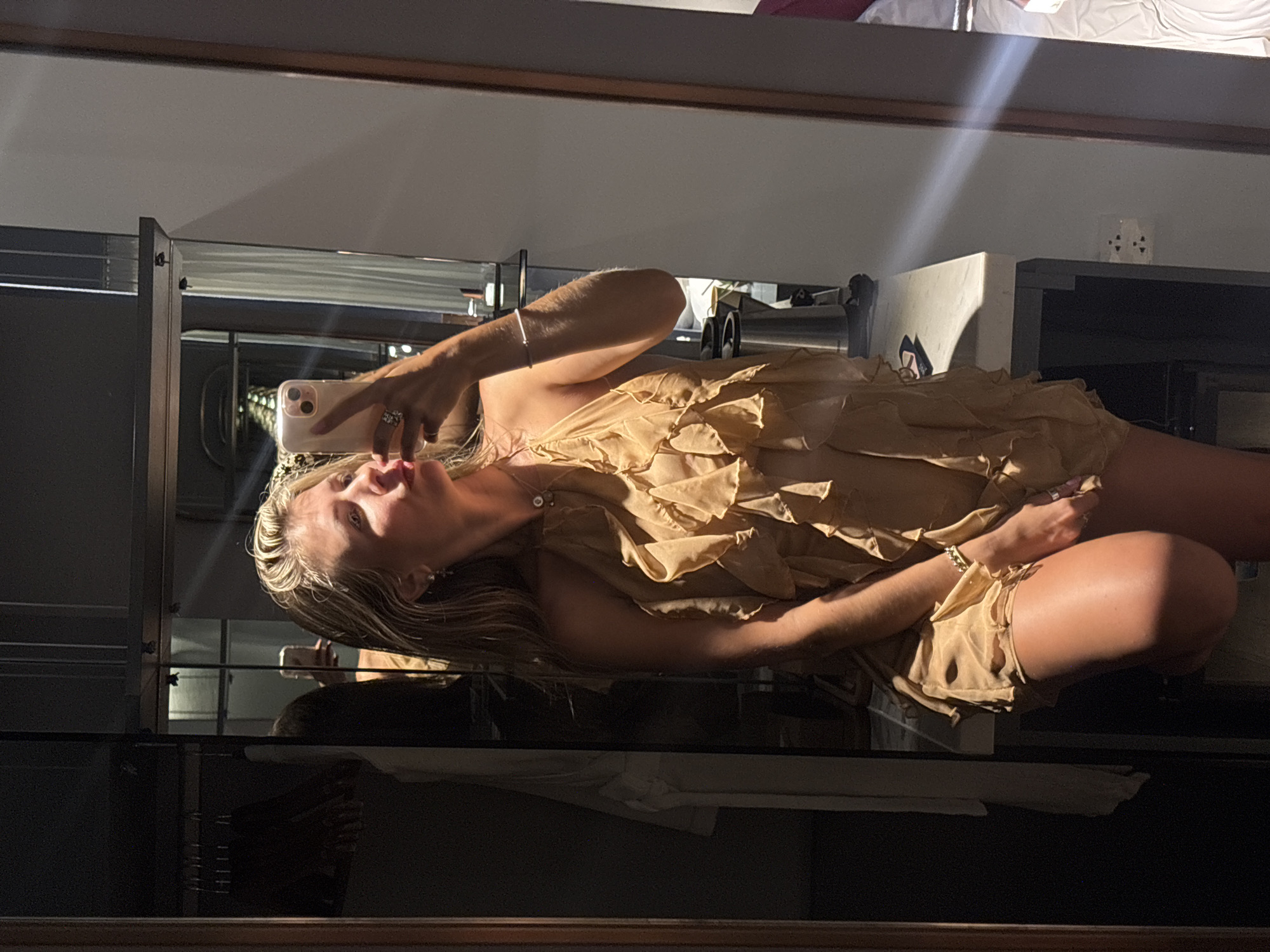 A young woman with blonde hair is taking a selfie in a hotel room. She is wearing a short, tan, ruffled dress and a silver bracelet. The woman is standing in front of a large mirror that reflects the room behind her, including a closet with clothes and a desk with a kettle and some papers. The lighting in the room is dim, with a strong beam of light hitting the woman from the side. The overall impression is one of a candid, intimate moment captured by the woman herself.