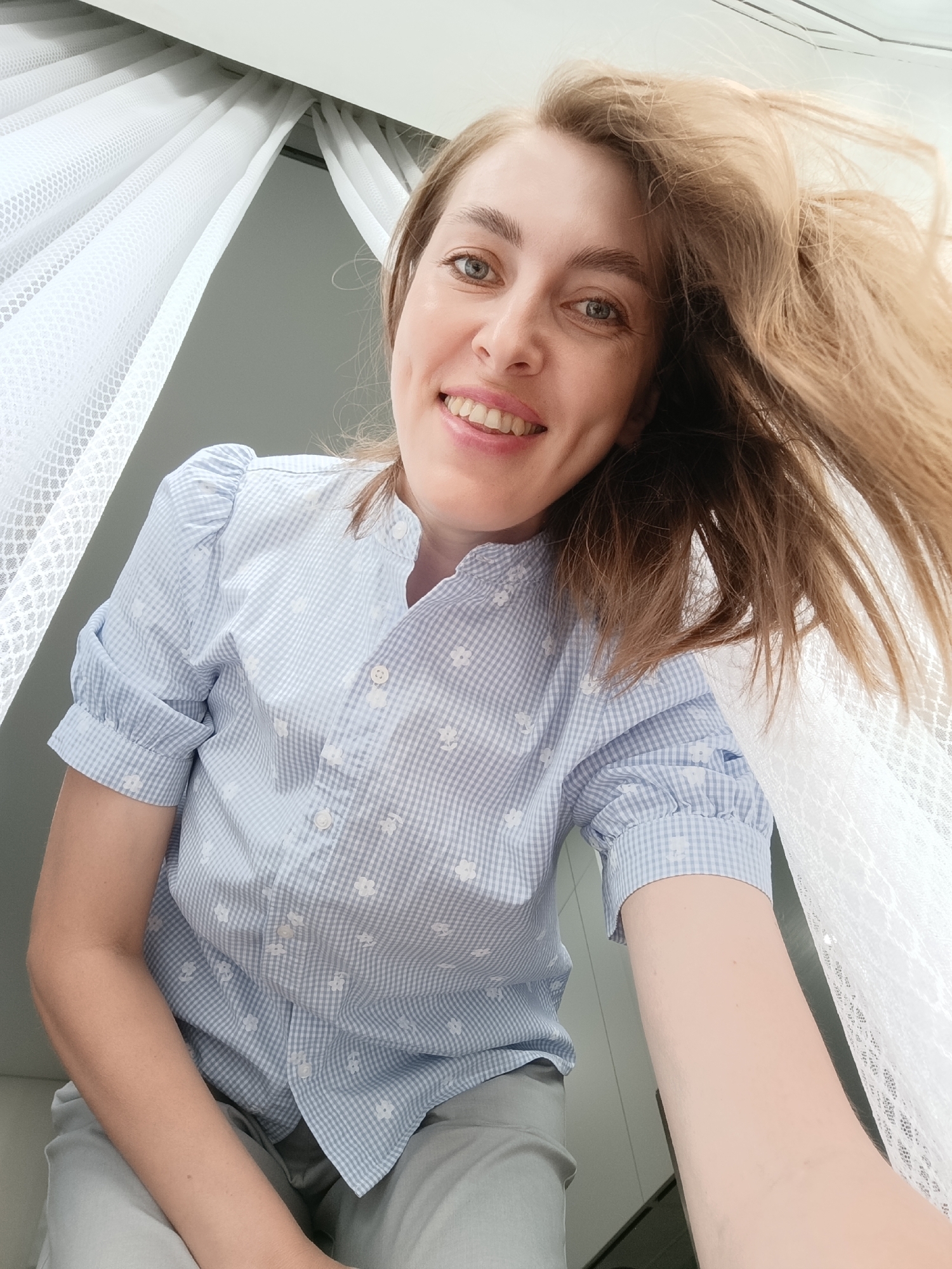 A woman is smiling into the camera. She is wearing a blue and white checkered shirt with short sleeves. Part of her light brown hair is up and to the right, blurring from movement. She appears to be indoors. Part of white, lace-like curtains are on the top left and bottom right corners of the image. The background is a light gray. There is no definitive evidence in the image to confirm it was taken in Chaikovsky, Russia.