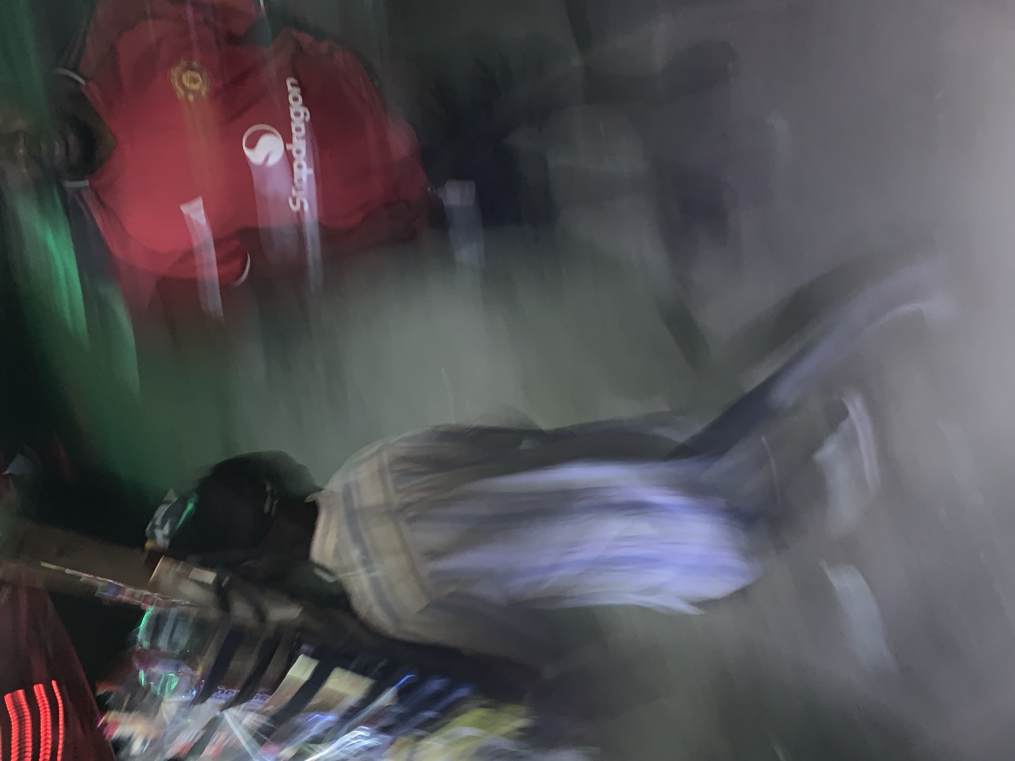 The image shows two people in a blurred and unclear setting, possibly taken with motion blur. One person is wearing a red soccer jersey with the word "Snapdragon" visible on the front. The jersey also appears to have the Manchester United logo. The other person is wearing what appears to be a striped shirt. The setting is dark and indistinct, with some lights visible in the lower portion of the image. There is no clear indication of the location being in New Stadium, Nigeria.