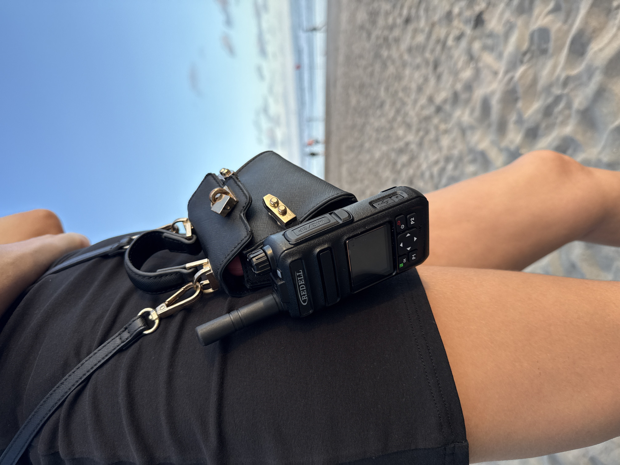 The image shows a close-up view of a person's lap while sitting on a sandy beach. The person is wearing a black outfit. A black purse with a gold-colored lock and a black walkie-talkie from the brand REDELL are placed on the lap. In the background, blurred figures of people can be seen on the beach, as well as the ocean and a blue sky. The location is North Kuta, Indonesia, based on the environment.