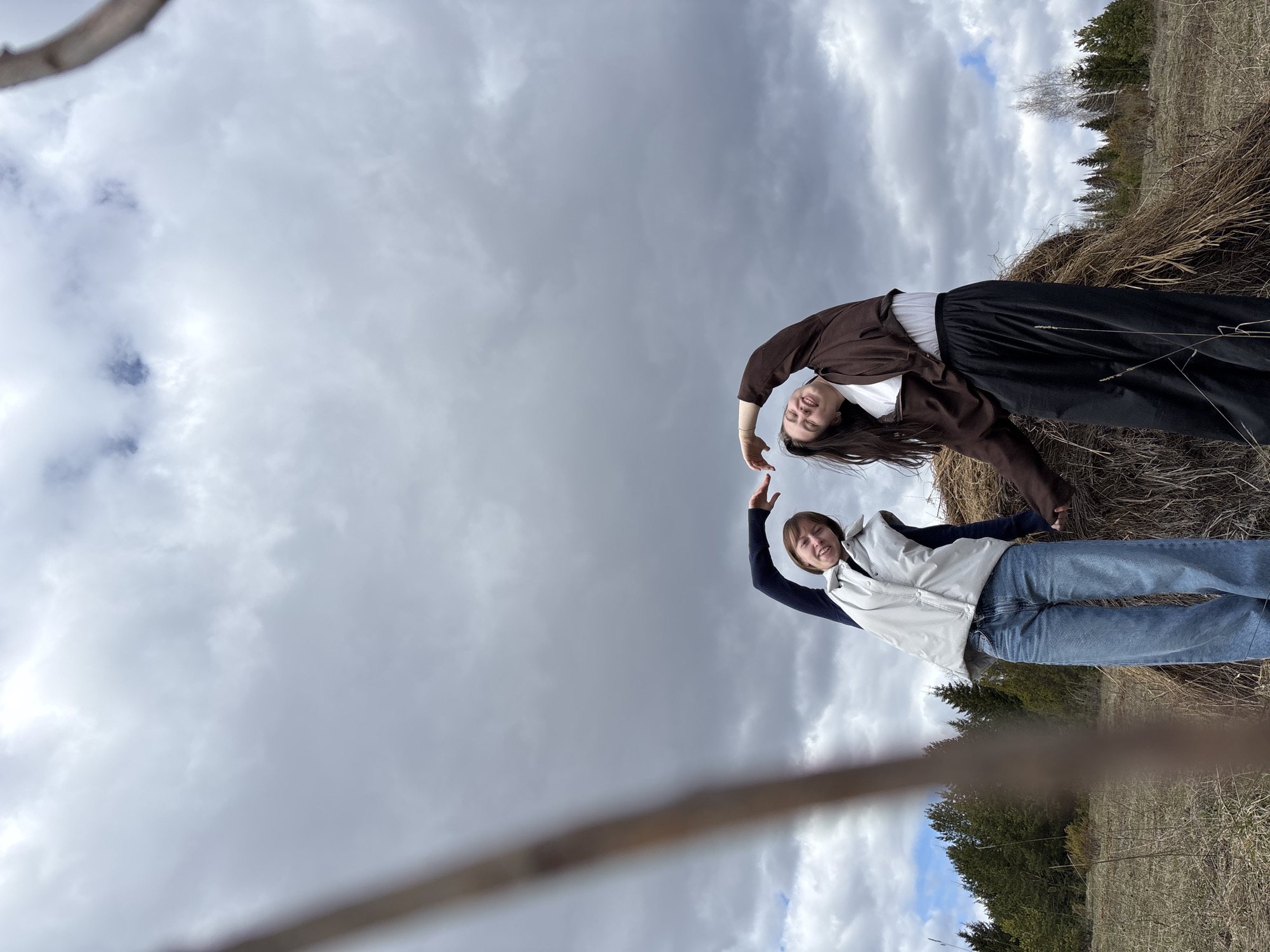 Two young women are posing outdoors under a cloudy sky. One woman is wearing a brown jacket, a white shirt, and black pants. She is leaning against what appears to be a haystack or a pile of dry grass. The other woman is wearing a dark long-sleeved shirt, a light-colored vest, and blue jeans. They are both smiling and reaching their hands towards each other as if forming a heart shape. The background includes fields, trees, and a cloudy sky. The specific location in the city of Петропавловск, Russia cannot be determined from the image alone.