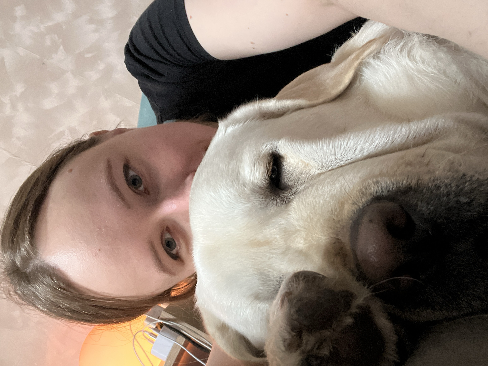 A woman and a dog are positioned closely, likely taking a selfie. The woman has fair skin, light brown hair, and light-colored eyes. She is wearing a black shirt. A white Labrador-type dog's head is nestled against her face. The dog's eyes are partially closed. The background appears to be a room with a textured wall. A charging device is visible in the lower left, along with an orange glow. No information indicates the image was taken in Mytishchi, Russia.
