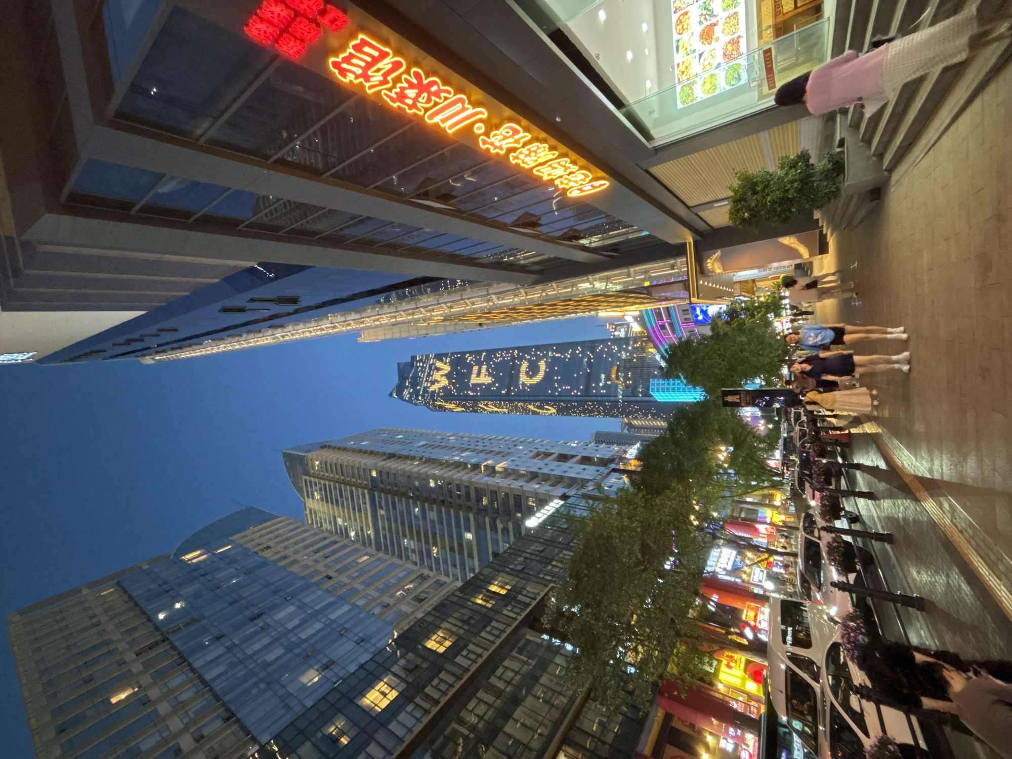This low-angle shot depicts a street scene at night in Yuzhong District, China, judging by the buildings and signage in view. Several tall, modern skyscrapers dominate the background, with one displaying the illuminated letters "BLU" vertically along its facade. The building to the top right displays glowing characters written in a Chinese script. Along the right side of the image, pedestrians walk on a brightly lit sidewalk, flanked by parked cars. Trees line the street, adding a touch of greenery to the urban environment. The sky is a deep blue, indicating either dusk or early evening. The perspective emphasizes the height of the buildings and the busy nature of the city.