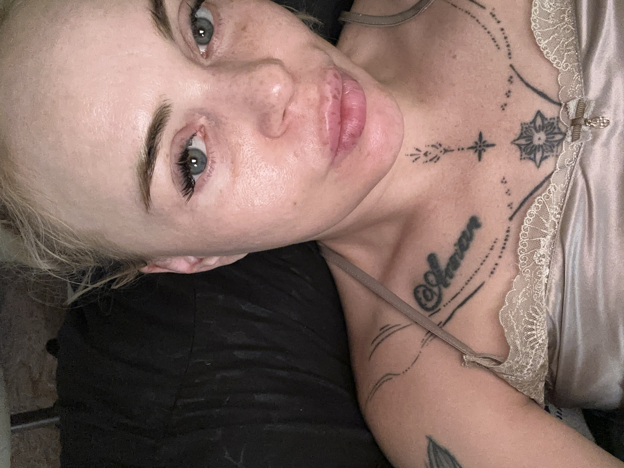 A close-up, overhead shot shows a fair-skinned person with blonde hair pulled back, lying down against a dark background. They are wearing a champagne-colored satin camisole with lace trim. Tattoos are visible on their chest, including the word "Amour" on the left side and a geometric design with a star in the center on the sternum. The person's face is looking directly at the camera, with eyes open and lips slightly parted. Their skin appears slightly red and shows some freckles. The lighting is dim and diffused.