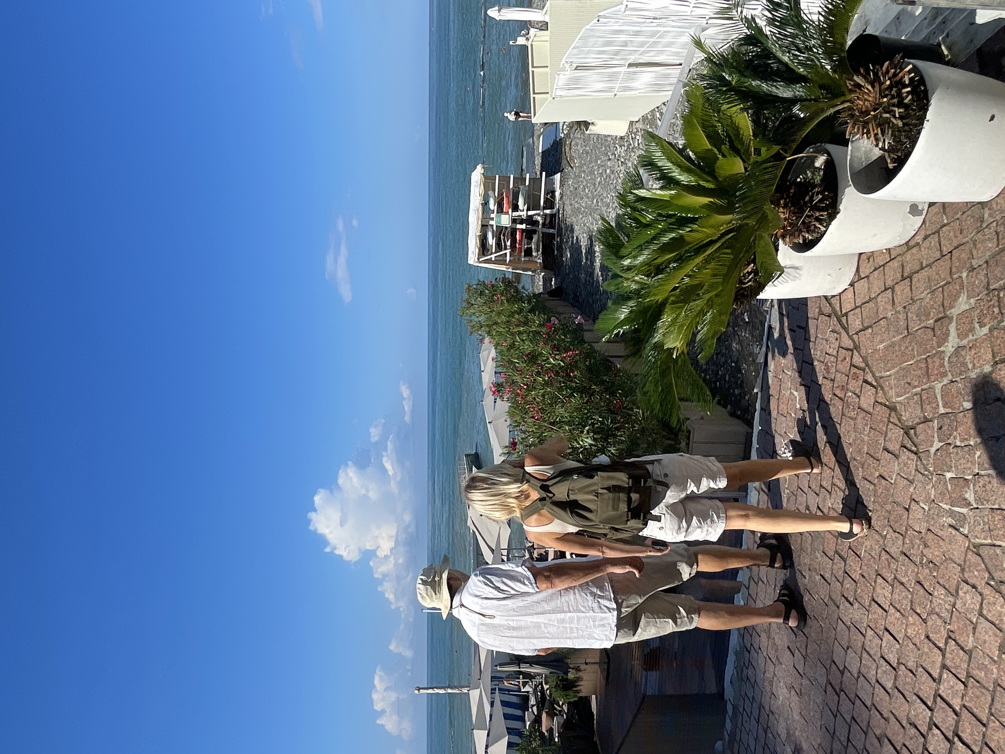 Two individuals, a man and a woman, are walking down a stone-paved pathway towards a beach scene in Sochi, Russia, on a sunny day. The man, wearing a light-colored short-sleeved shirt, shorts, and a hat, is on the left. The woman, in a white tank top and shorts, is on the right and carries a green backpack. They are facing away from the camera, looking towards the sea.
The background reveals a beach with rows of umbrellas, lounge chairs, and a structure holding several surfboards or paddleboards. The sea is calm with a few people visible in the water. Lush green plants in white planters are positioned on the right side of the pathway. The sky is a clear blue with scattered white clouds. The scene captures a relaxed, resort-like atmosphere characteristic of Sochi's coastal areas.