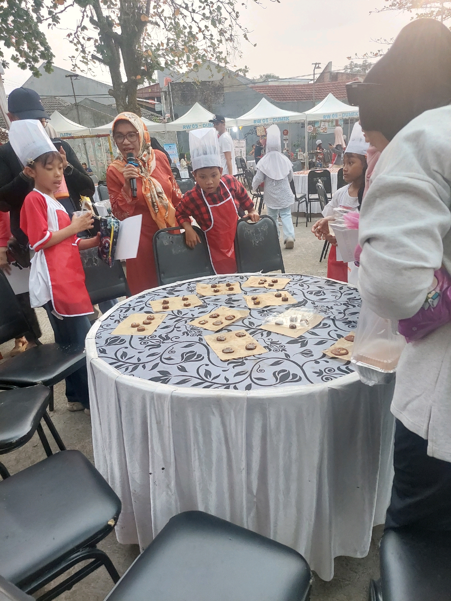 In Depok, Indonesia, a group of children and adults are gathered around a round table covered with a white tablecloth decorated with a black floral pattern. The table displays numerous small, brown, round pastries, each individually placed on pieces of brown paper. Several children, wearing chef hats and aprons, are present. One child in a red and white apron holds a clear plastic cup and a piece of paper. A woman wearing a headscarf and glasses is speaking into a microphone, facing the children. In the background, tents with the inscription "RW 05 - DUMEK" are visible, suggesting an outdoor event or fair. Several black chairs surround the table. The overall scene appears to be an event involving children, possibly a cooking or baking activity, taking place outdoors in a semi-urban environment.