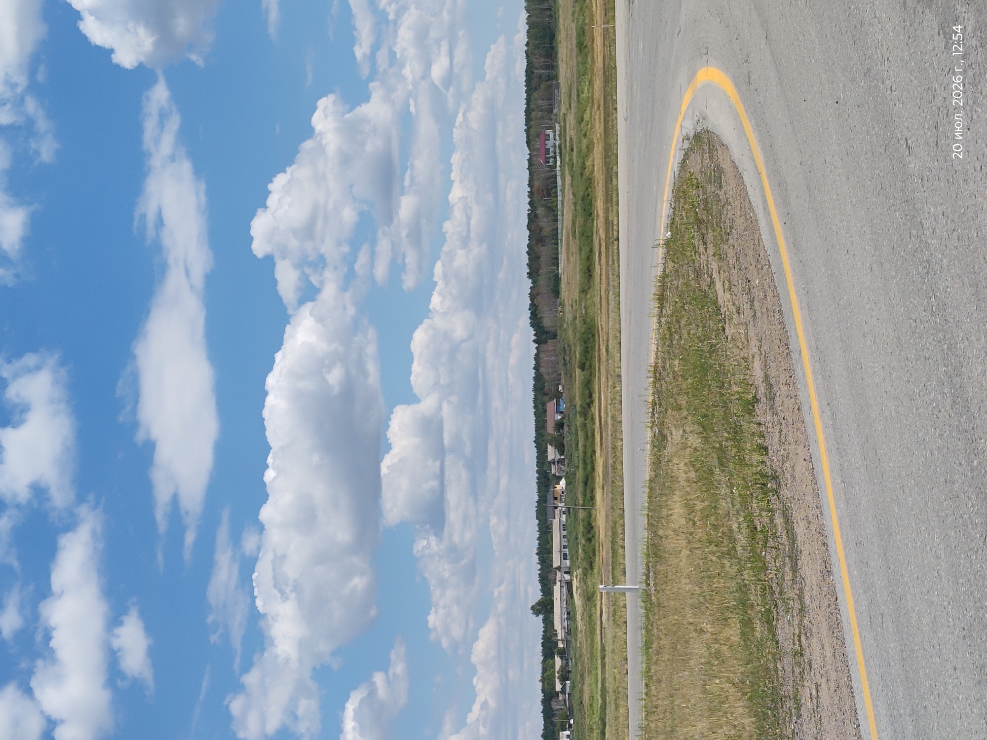 The image shows a rural landscape under a partly cloudy sky. In the foreground, a curved asphalt road with a yellow line marking its edge is visible. Dry grass and some sparse vegetation line the side of the road. In the middle ground, a cluster of low-rise buildings, possibly a village, is situated behind a strip of green fields. Further in the background, a dense line of trees forms a treeline, and beyond that, the horizon is visible under a bright blue sky filled with white, fluffy clouds. The overall scene depicts a quiet, open area, characteristic of a rural setting in a location such as Айдарлы, Kazakhstan. There are no people or animals visible in the image. The date stamp at the bottom right corner indicates "20 июл. 2026 г., 12:54".