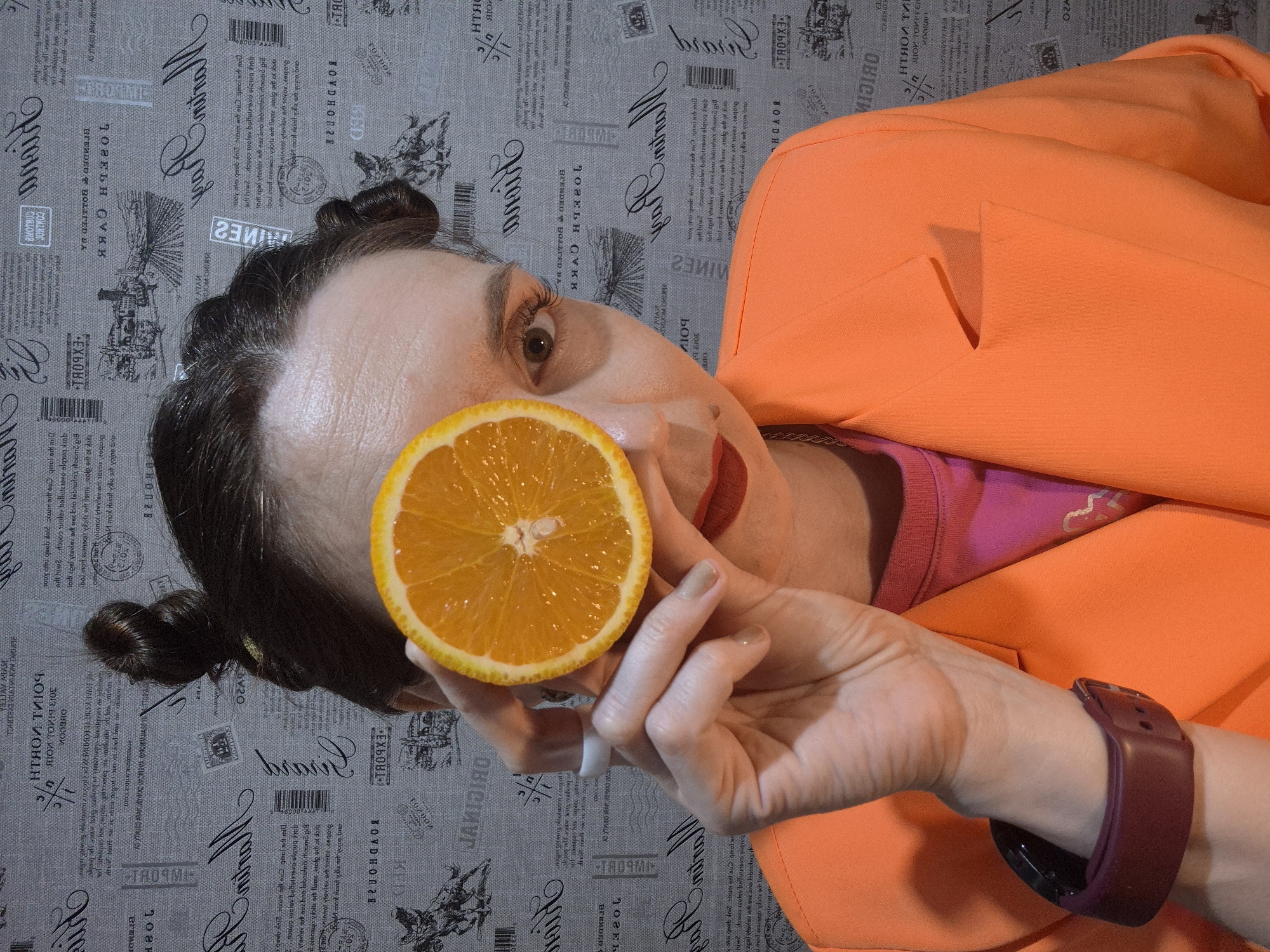 A woman with dark hair styled in rolls is holding a half-cut orange in front of her eye. She is wearing an orange blazer over a pink and burgundy top. A burgundy smartwatch is visible on her wrist. The background is a wallpaper with newspaper print.