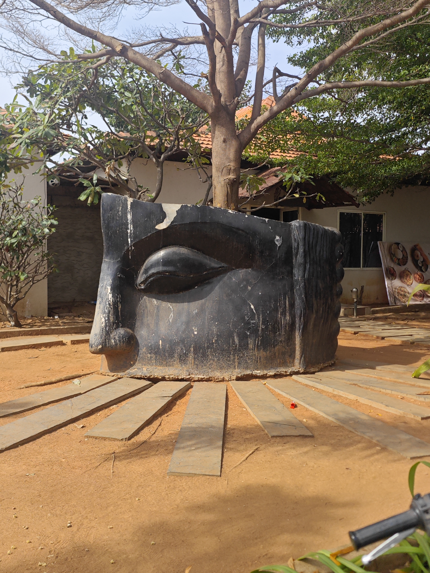 A large, black, weathered statue of a face, possibly depicting a deity or an artistic representation, is centrally located in an outdoor area with sandy, orange-brown ground. The statue is partially submerged in the ground and surrounded by a radial pattern of rectangular, flat stone or concrete slabs. Behind the statue, a building with a tiled roof and white walls is visible, with trees and foliage partially obscuring it. The trees have both bare branches and green leaves, suggesting a possibly temperate climate. In the background, a large poster displaying various food items on plates is leaning against the building. The image appears to be taken during the daytime, with shadows cast by the trees and the statue. The location is identified as Aruru, India.