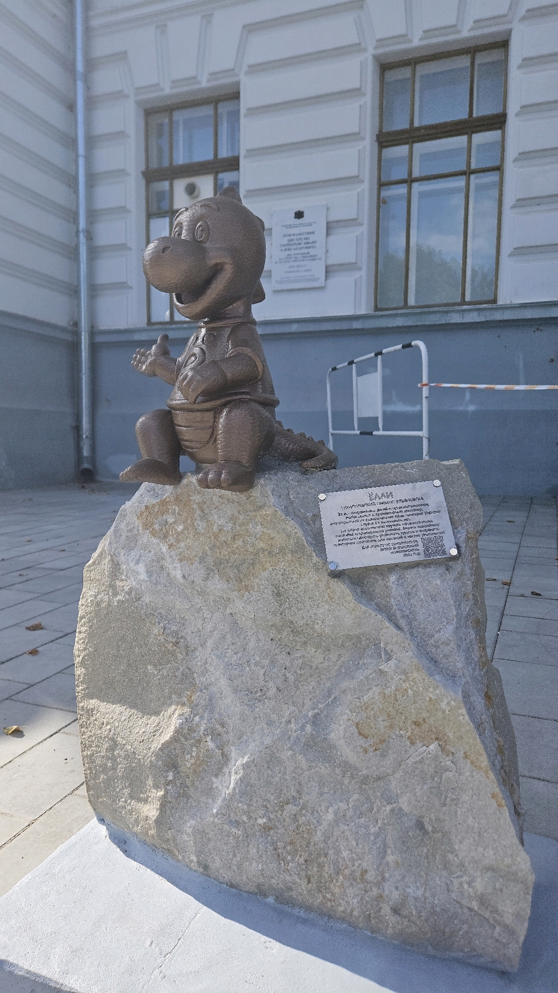 A bronze sculpture of a cartoonish dragon character, possibly a mascot, is situated outdoors on a large, rough-hewn light-colored rock. The dragon is depicted in a seated position with its arms outstretched and hands open, facing slightly to the left. It has a wide grin and large, expressive eyes. The sculpture is dark bronze in color. Attached to the front of the rock is a silver plaque with text and a QR code. Behind the rock and sculpture, a white building with classical architectural elements, including arched windows and decorative molding, is visible. A metal railing with white and orange tape is also present in the background. The ground in front of the rock is paved with square tiles. The scene is illuminated by daylight, casting shadows.
This is a photograph of the "Dragon Klava" sculpture located on the embankment of the Volga River in Ulyanovsk, Russia.