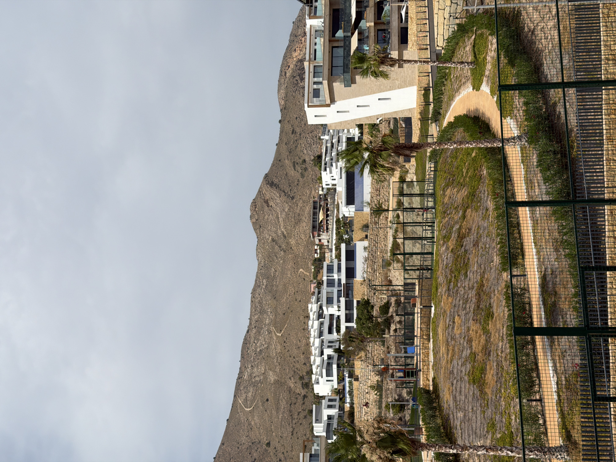 This is a medium shot of a modern residential complex in Finestrat, Spain. In the foreground, a dark green metal fence with a diamond pattern encloses a tan-colored path. Beyond the fence, to the left, is a palm tree with its fronds reaching upwards. To the right, a second palm tree stands similarly. A beige path curves through the landscape, bordered by low-lying green shrubs and scattered patches of dry grass and dirt. In the middle ground, a red and blue playground structure is visible, enclosed by a fence. Further back, a series of white, cube-shaped buildings with dark windows are terraced up a hillside, topped by a larger, angular white building with multiple balconies and glass railings. The buildings are set against a backdrop of a brown, rocky hillside with a winding path visible. The sky is overcast and light grey.