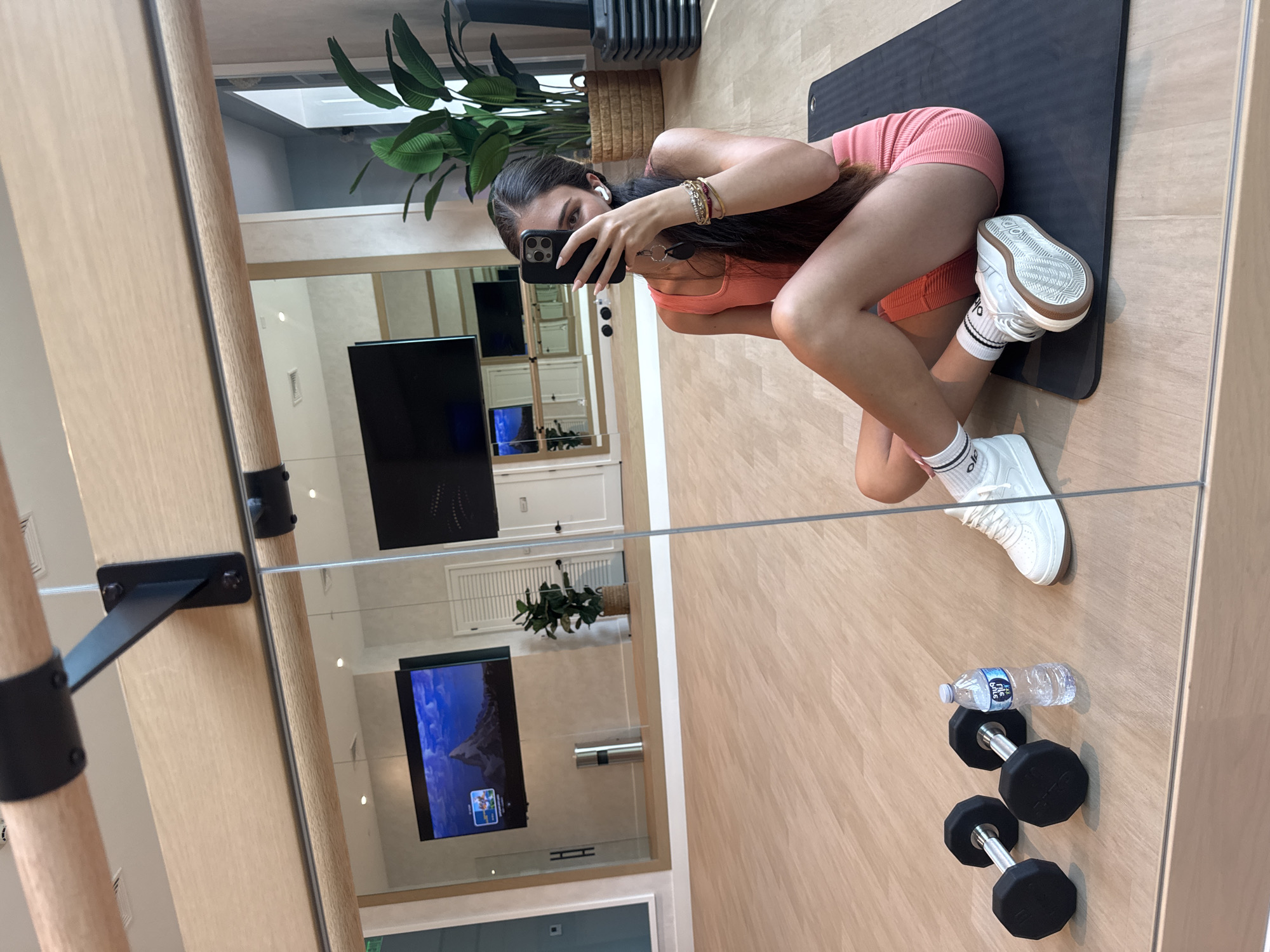 A young woman with long, dark hair is taking a mirror selfie in a gym. She is wearing a coral-colored tank top and matching shorts, white socks with black stripes, and white sneakers. She is sitting on a black yoga mat with a bottle of water and two dumbbells to her left. The gym has light wood floors and mirrors on the walls. In the background, a large television screen displays a mountain landscape, and a potted plant sits in the corner. Other gym equipment, including step platforms, is visible in the reflection. The woman is holding her phone up to take the photo, and her other hand is resting on her leg, adorned with several bracelets. She has white earbuds in her ears.