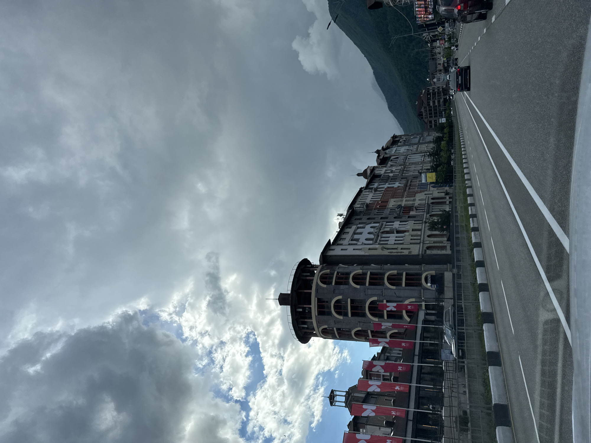 This is a vertical, eye-level, medium shot of a street scene in Estosadok, Russia, under a cloudy sky. The road in the foreground has visible white lane markings. Several cars are on the road, moving in the same direction. On the left side of the road, there are several buildings with multi-colored facades, some featuring red banners with white lettering. A prominent cylindrical tower with arched windows stands out among the buildings. To the right of the buildings, a lush green mountainside rises, partially obscured by clouds. The overall atmosphere is overcast.