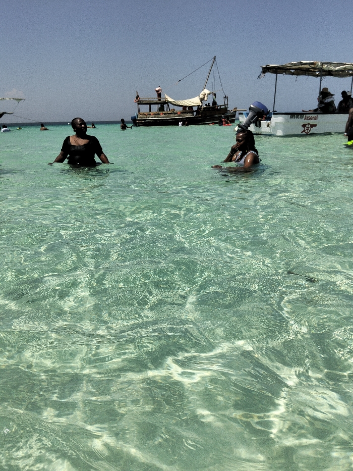 This image depicts a marine leisure scene in Wasini, Kenya, under a clear blue sky. The foreground and midground feature clear, shallow, light turquoise-green water.
In the midground, two dark-skinned females are partially submerged. The woman on the left stands chest-deep, wearing a dark short-sleeved top, facing slightly right with her hands near her hips. The woman on the right sits chest-deep, wearing a light-colored top, facing slightly left, with her hands near her chest, possibly interacting with an object or communicating.
In the background, a traditional wooden dhow boat with a mast and a partially furled light-colored sail is visible to the left. Several individuals are on board or alongside the dhow. To the right, a white fiberglass speedboat with a blue outboard motor and a canopy is present. Text on its hull includes "ARSENAL" and a partial registration number, possibly "KW 0725H". Multiple individuals are seated within this speedboat. Additional people are visible in the water and near other smaller vessels in the far background. The scene suggests a sunny day with recreational activity in the coastal waters.