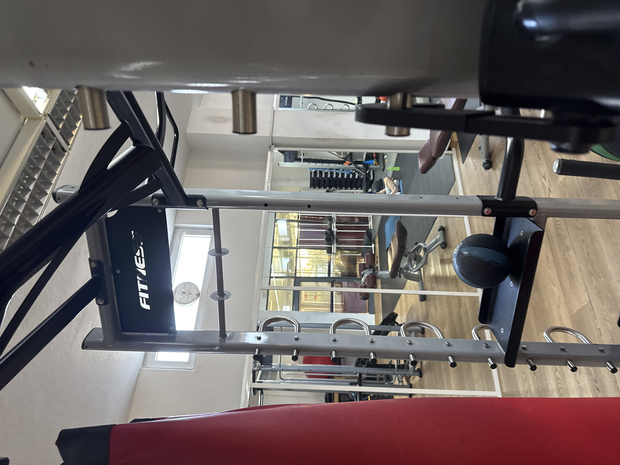 The image shows an indoor gym environment, likely in Mirandela, Portugal based on the geotag. The perspective is from behind gym equipment, specifically a power rack or similar fitness apparatus. Parts of the gray metal frame of the rack are prominently in the foreground. A black panel with the white text "FITNESS" is visible on one of the frame members. In the background, mirrored surfaces reflect additional gym equipment, including weight benches and a squat rack, along with a person lying on the floor. A black and blue medicine ball rests on a platform attached to the rack. The floor appears to be a light-colored wood or laminate. A red pad or piece of equipment is partially visible in the foreground.