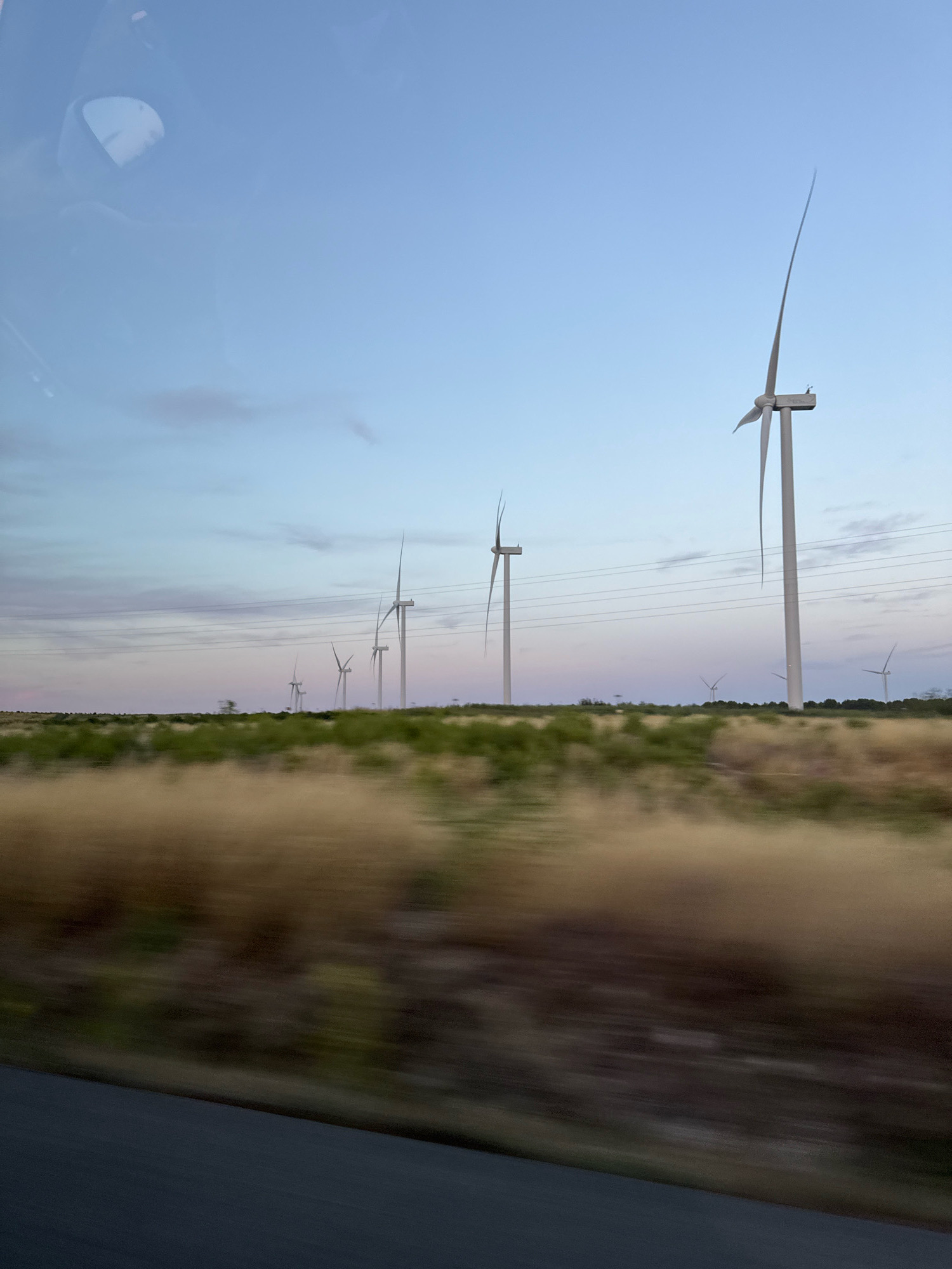 A line of wind turbines stretches across a rural landscape under a twilight sky. The foreground features a blurred expanse of dry grass and vegetation, suggesting motion. In the middle ground, a line of green bushes is visible, bordering a flat, open area where the wind turbines are situated. The turbines themselves are tall, white structures with blades, some of which are in motion. Power lines are strung between poles in the distance. The sky transitions from a pale blue at the top to hues of pink and purple near the horizon, indicating dawn or dusk. A reflection of a car's side mirror is visible in the upper left corner of the image, implying the photo was taken from inside a vehicle. The overall scene depicts a wind farm in a rural setting, likely in Cadreita, Spain, during a transitional period of daylight.