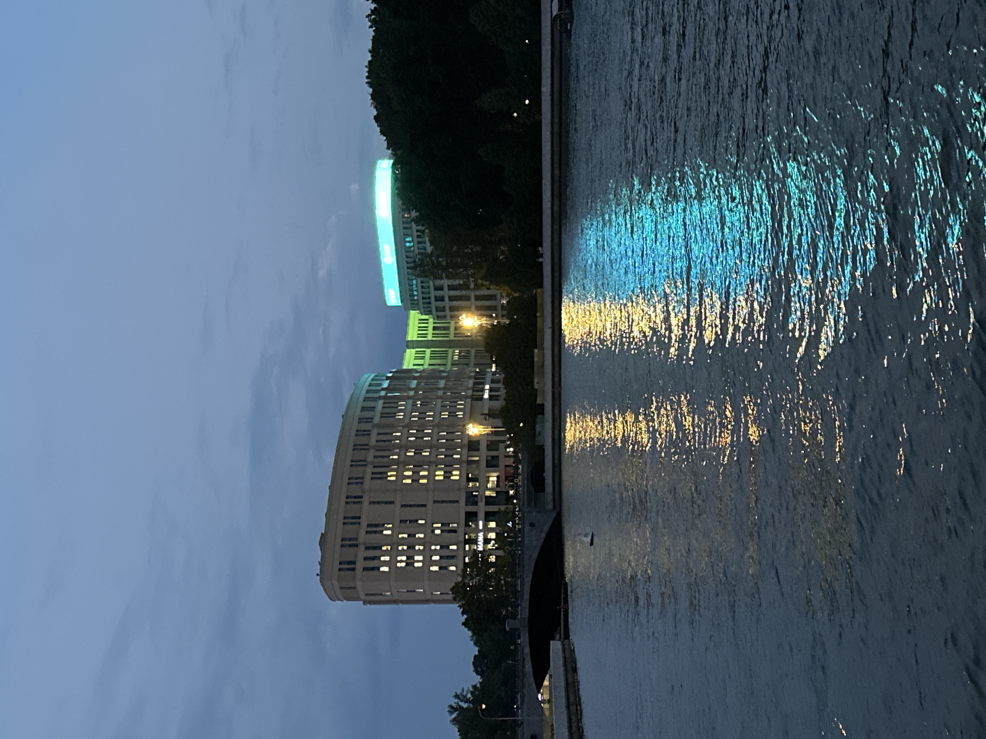 The image captures an evening view of the Svislach River in Minsk, Belarus, with two modern buildings illuminated on the far bank. The sky is overcast and a deep blue, indicating dusk. The river occupies the foreground, its surface reflecting the lights from the buildings and the sky.
The building on the left is a curved, multi-story structure with many windows, some of which are lit with a warm yellow light. The building on the right is also multi-story and features a prominent, brightly lit sign in a vibrant turquoise and green color scheme on its upper section. A bridge arches over the river in the middle ground, connecting the riverbanks. Dark trees line the riverbank behind the buildings. A small white sailboat with a red hull is visible on the water, near the bridge. The overall scene is tranquil and picturesque, with the reflections of the lights creating a shimmering effect on the water.