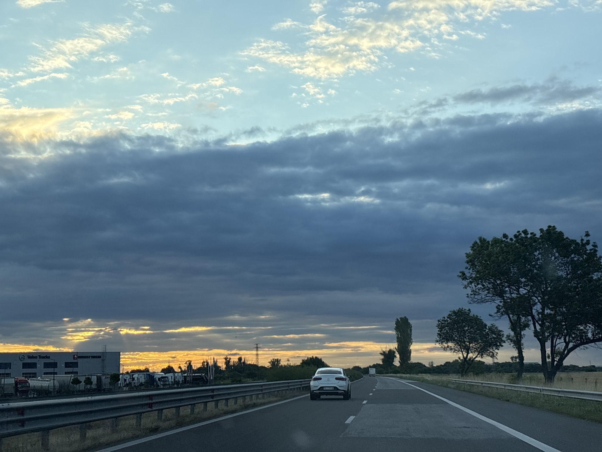 The image depicts a view from a vehicle traveling on a highway, presumably near Benkovski, Bulgaria. The sky is a prominent feature, with layers of clouds, some dark and heavy, others lighter and tinged with yellow from the setting or rising sun. Shafts of light break through the darker clouds on the left side of the frame, illuminating the horizon.
On the left side of the highway, a large industrial building is visible, with the text "Volvo Trucks" clearly displayed on its facade. Several trucks and trailers are parked in front of the building. The highway itself is paved with asphalt, marked with white lines indicating lanes. A metal guardrail runs along the side of the road.
In the foreground, a white car is driving ahead on the highway. Further down the road, more vehicles and trees are visible. The landscape on the right side of the highway consists of fields and trees, including several tall, slender trees and a larger, fuller-crowned tree. The overall impression is a highway scene at dusk or dawn, with dramatic cloud formations and industrial elements on one side, and natural landscape on the other.