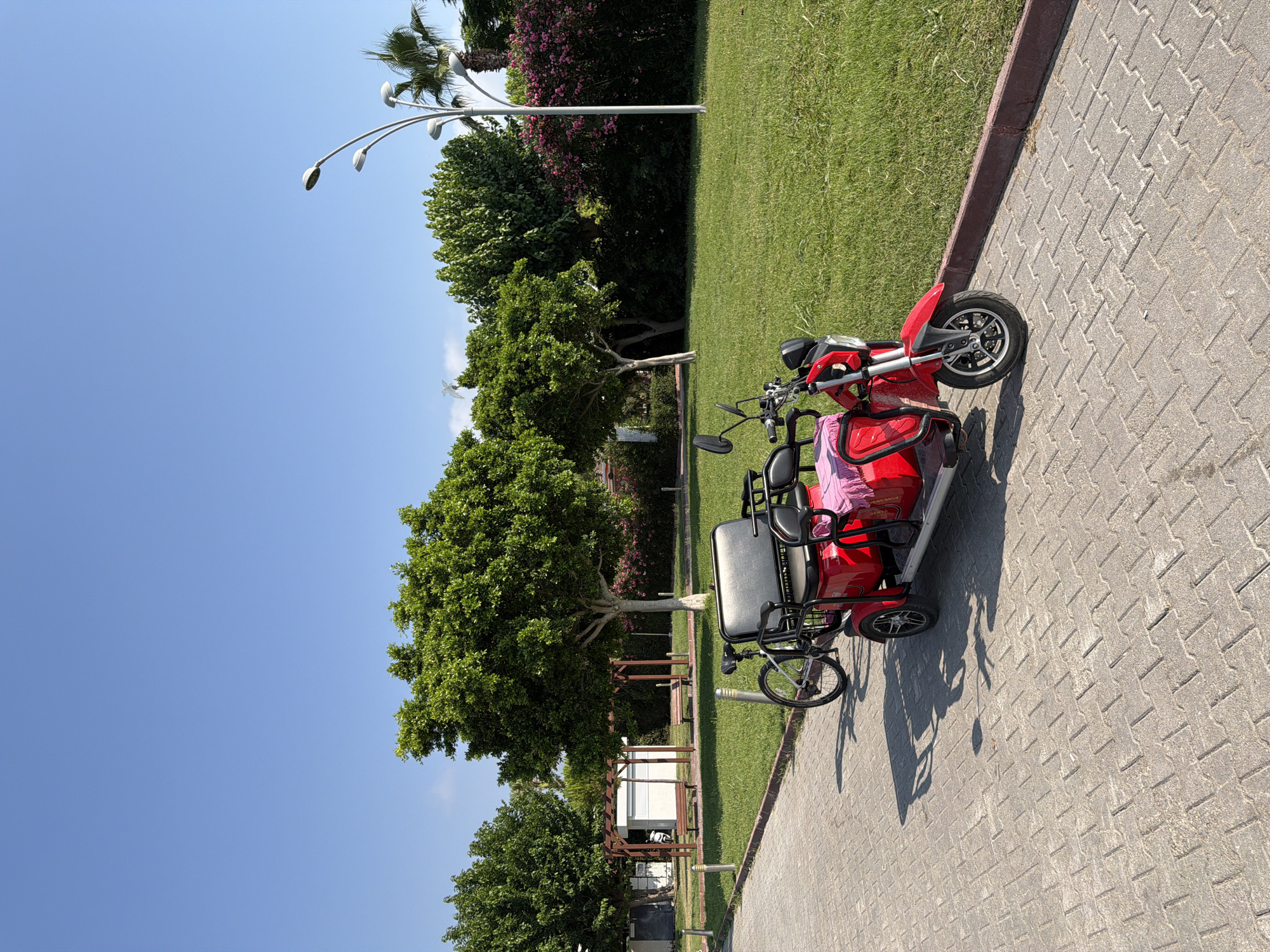 In Kemer, Turkey, on a sunny day, a red electric tricycle with two seats and a pink blanket draped over it is parked on a paved walkway. A bicycle is positioned behind the tricycle, its front wheel aligned with the tricycle's rear wheel. The walkway is bordered by a strip of grass, beyond which a lush green lawn stretches, dotted with trees and flowering bushes. A modern lamppost stands on the right side of the lawn, its lights pointing upwards. In the background, a wooden pergola with benches under it is visible, suggesting a recreational area. The sky above is a clear, bright blue.