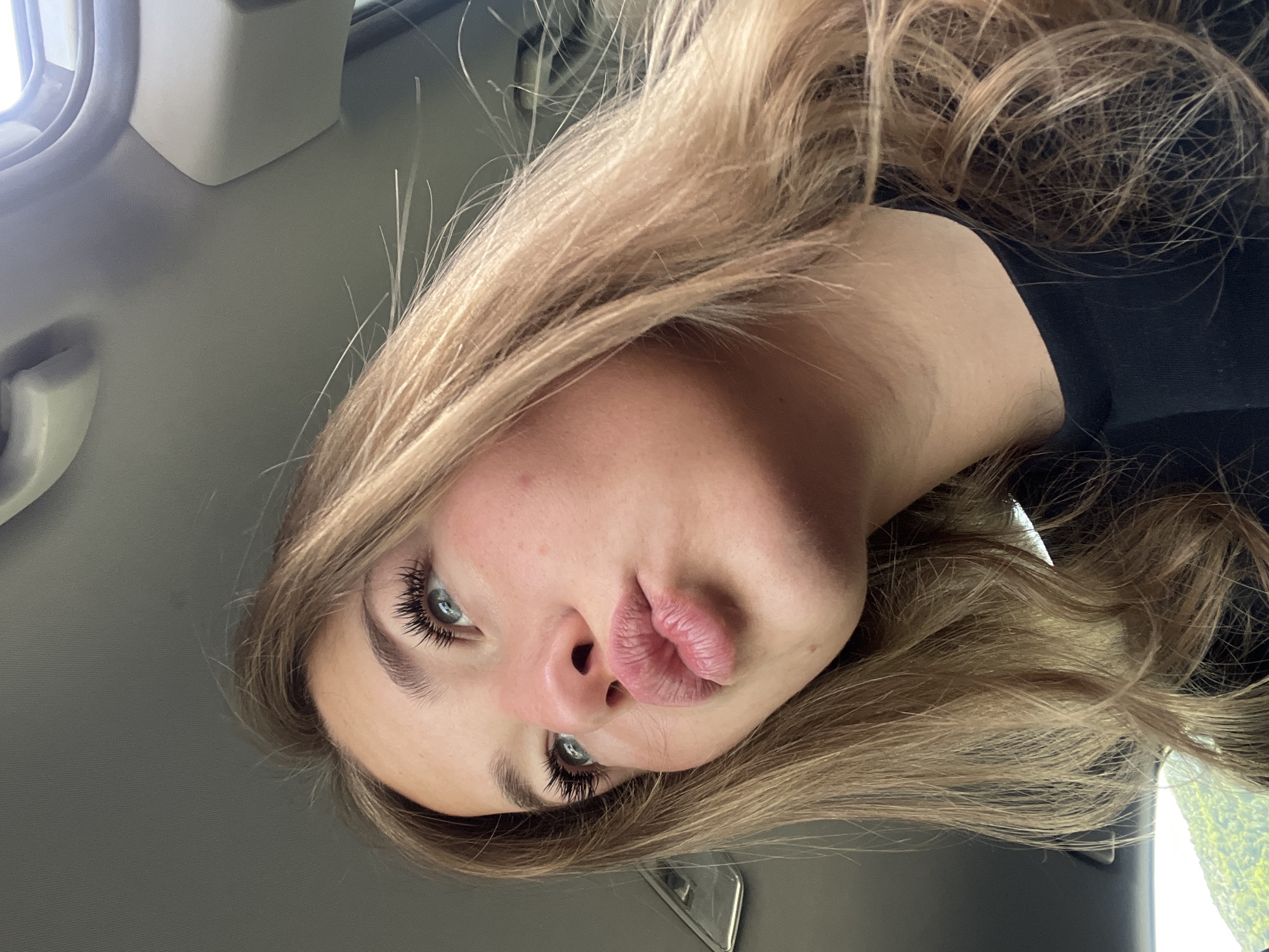 A young woman is sitting in a car. She has long blonde hair and blue eyes. She is wearing a black shirt. She is making a kissing face towards the camera. The car interior is gray. The location is not determinable from the image.