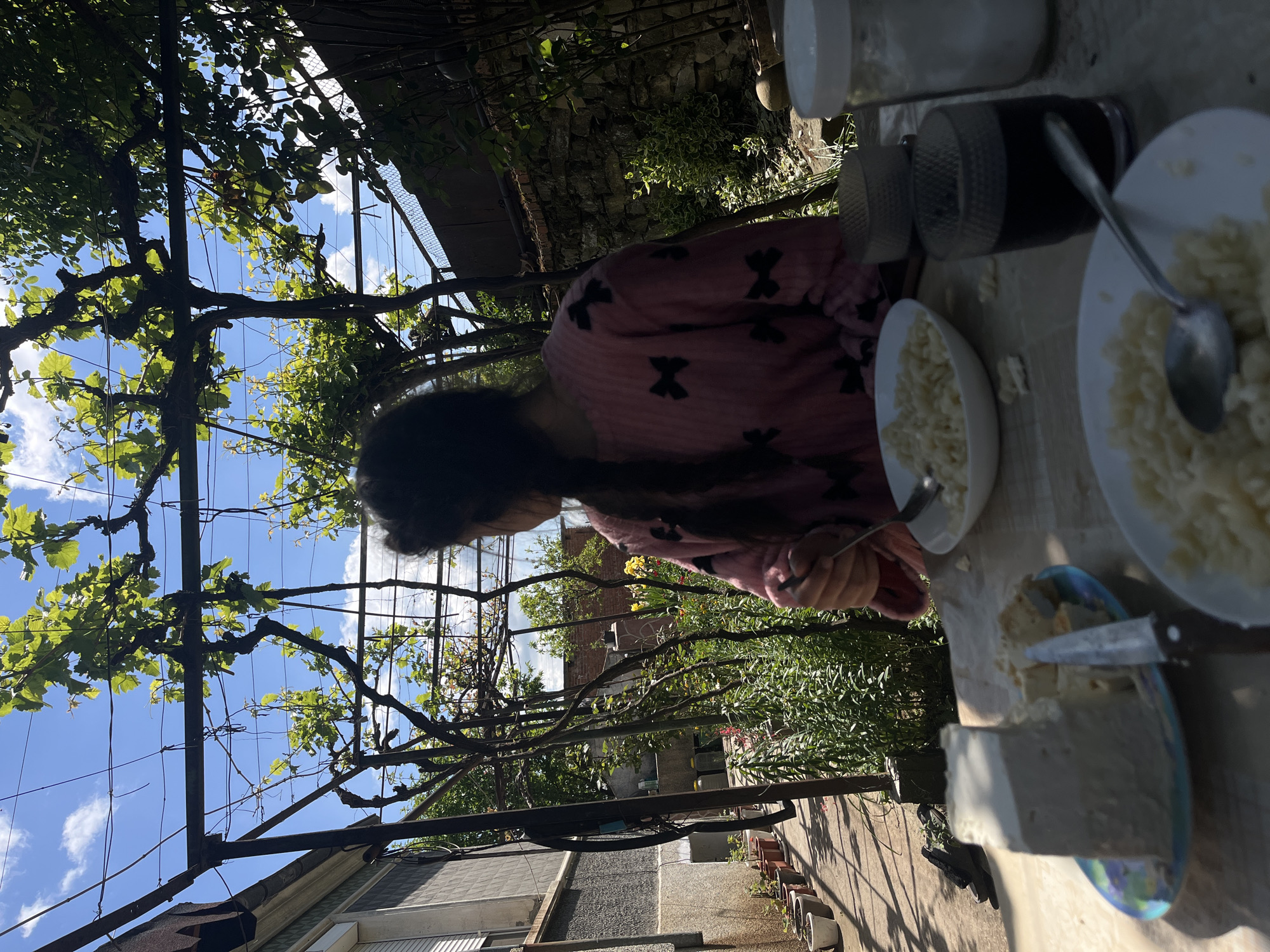 A person with dark hair is sitting at a table outdoors. The person is wearing a pink robe with black crosses on it. On the table are plates of cooked pasta, and cheese. The table is in a garden or yard, with trees and bushes visible in the background. A vine-covered pergola structure is overhead. The location is inferred to be in Lozarevo, Bulgaria.