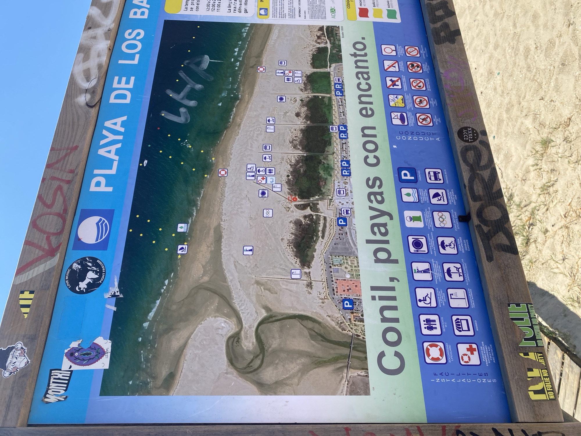 The image depicts a large informational sign located on a beach in Conil de la Frontera, Spain. The sign is mounted on a wooden frame and is primarily blue and green. At the top, the sign displays "PLAYA DE LOS BA" in white lettering, suggesting it is part of a title related to a beach. Below this, a map of the beach area is visible, showing the coastline, sandy areas, and some vegetation. Several small icons are scattered across the map, indicating various amenities or zones.
The lower portion of the sign features text and icons. A large green band across the bottom reads "Conil, playas con encanto." Below this, there are two columns of icons with corresponding text in Spanish. The left column, labeled "INSTALACIONES FACILIDADES," includes icons for a lifeguard, restrooms, accessible restrooms, controlled beach access, food and drink service, and sports zones. The right column, labeled "CONDUCTA," displays icons related to beach regulations, such as no campfires, no horses, no dogs, no tents, and no vehicles.
The sign itself shows signs of wear and tear, with stickers and graffiti applied to it. Notably, there are several stickers with graphics and text, including one with a monkey-like figure and the word "MONTHI." Graffiti in red and white spray paint is visible on the wooden frame and the blue section of the sign. The background of the image shows a sandy beach with the ocean in the distance, indicating it is an outdoor, sunny environment. The overall scene is an informative public display on a beach, subject to the elements and public interaction.