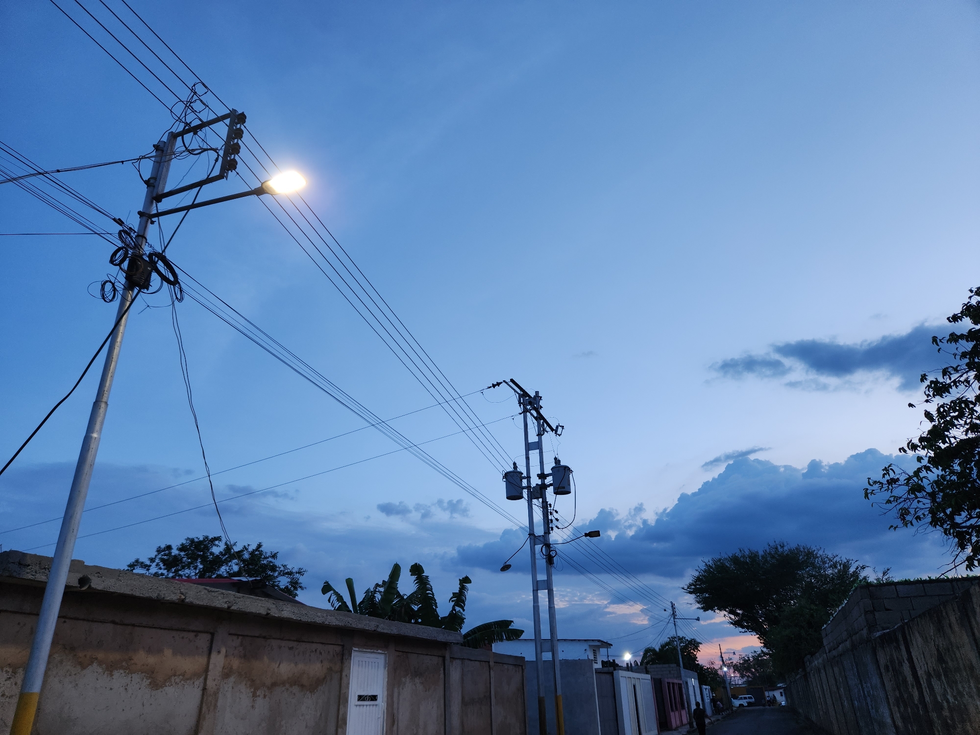 The image depicts a street scene in Tinaquillo, Venezuela, during twilight. The sky is a gradient of blue, with darker clouds in the distance. Two utility poles with electrical transformers and a street light are visible. The street is flanked by walls and buildings of varying heights and conditions. Some vegetation is visible, including trees and banana plants. A person walks in the distance down the street.