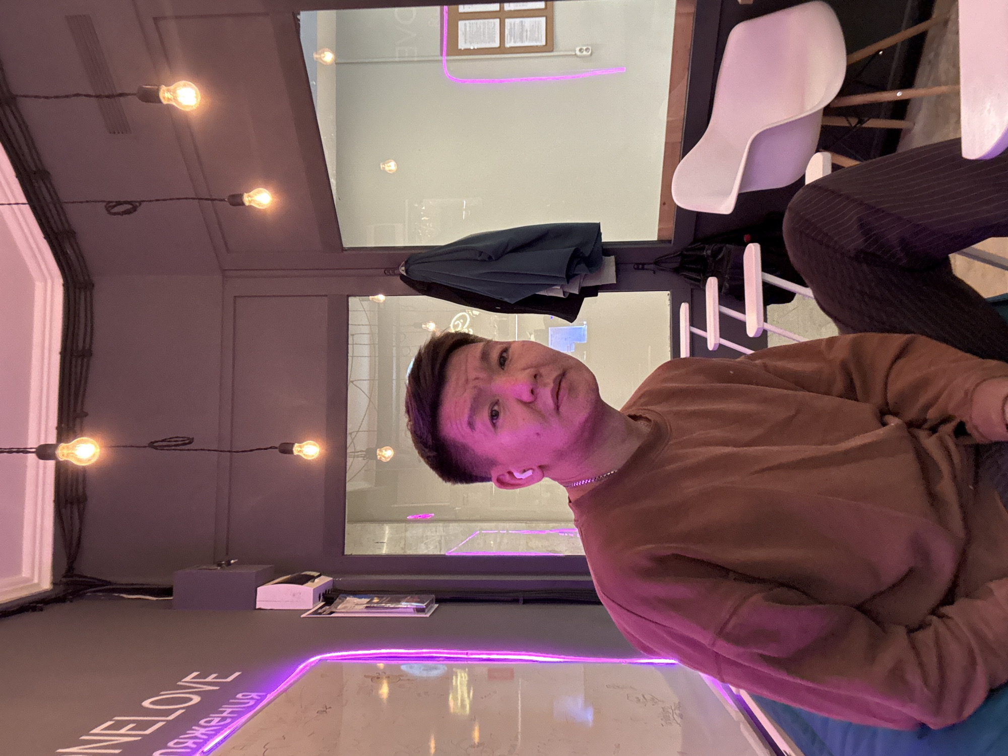 A medium eye-level shot captures a man in a brown sweatshirt indoors. He has dark hair and is wearing a silver chain. He is sitting next to someone wearing striped pants. The backdrop features a blurred cityscape visible through a window, lights hanging from the ceiling, and a pink neon sign. A white chair is visible near the window. The text "NELOVE" is illuminated in pink neon on a table. The location is Moscow, Russia.