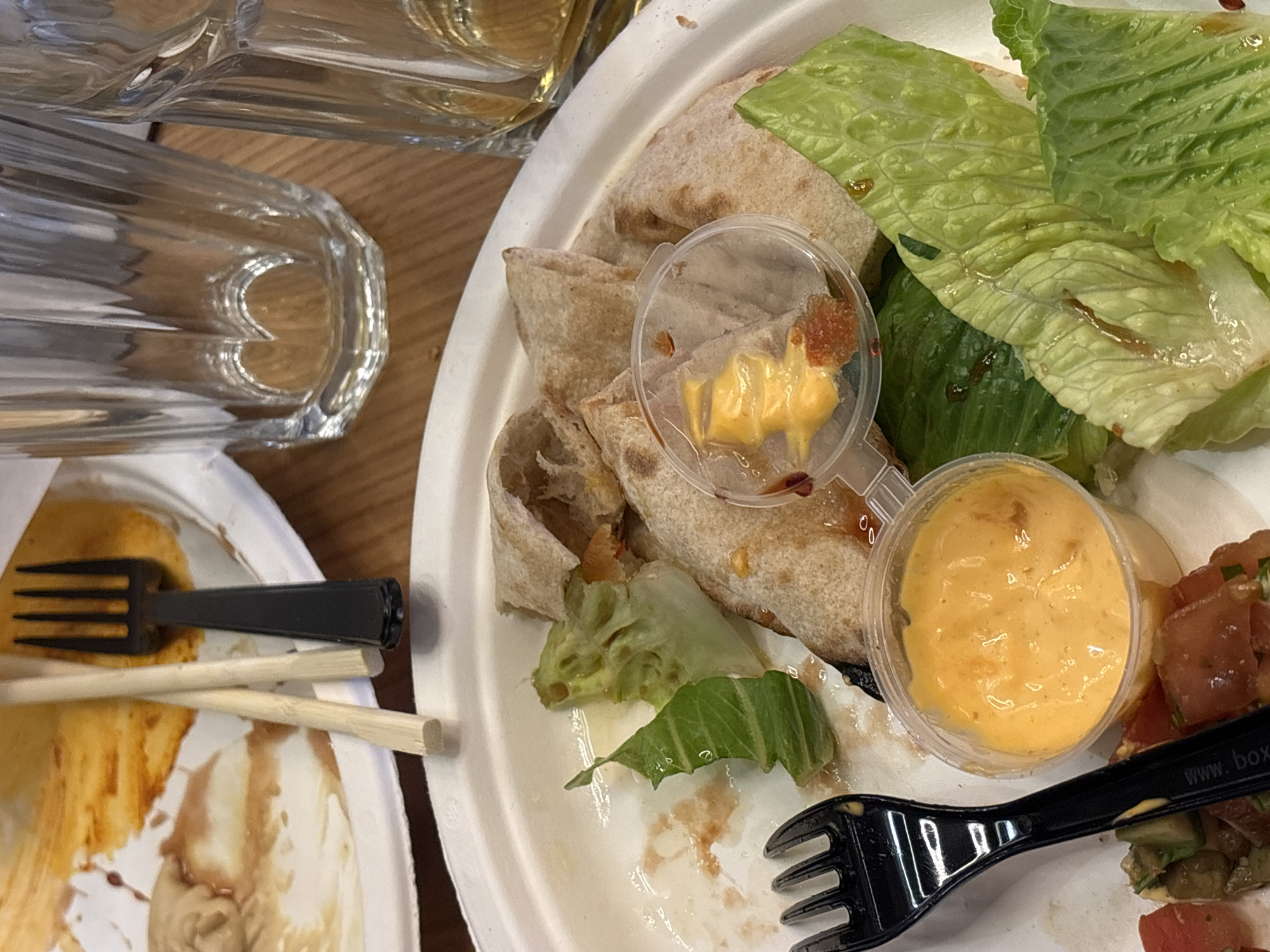 The image shows a partially eaten meal on a white disposable plate. The meal appears to be a wrap or burrito, cut into pieces, accompanied by lettuce, tomato salsa, and two small plastic containers of yellow sauce. One of the sauce containers has already been used. The plate is placed on a wooden surface. To the left of the plate, there's another disposable plate with leftover food, a black plastic fork, and a pair of wooden chopsticks. A glass container, possibly a water pitcher, is partially visible in the upper left corner. The food appears to be consumed. It is not possible to determine if the location is in Moscow, Russia, or to describe people or interactions from this image.