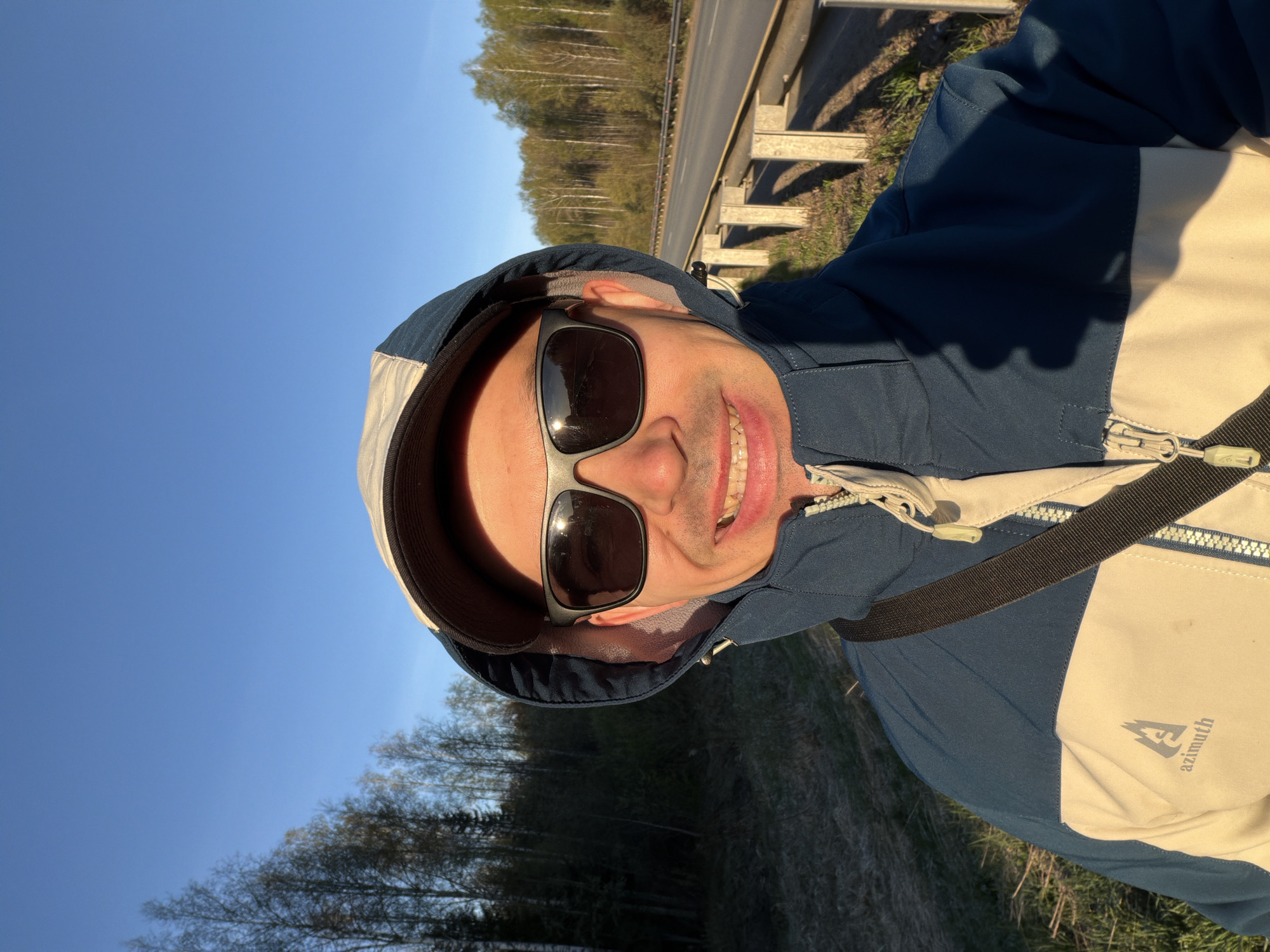 A man is taking a selfie outdoors. He is wearing sunglasses and a two-tone blue and beige hooded jacket with a strap across his chest. He is smiling and standing near a road with a guardrail. A forest is visible in the background under a clear blue sky. The location is Люкшудья, Russia.