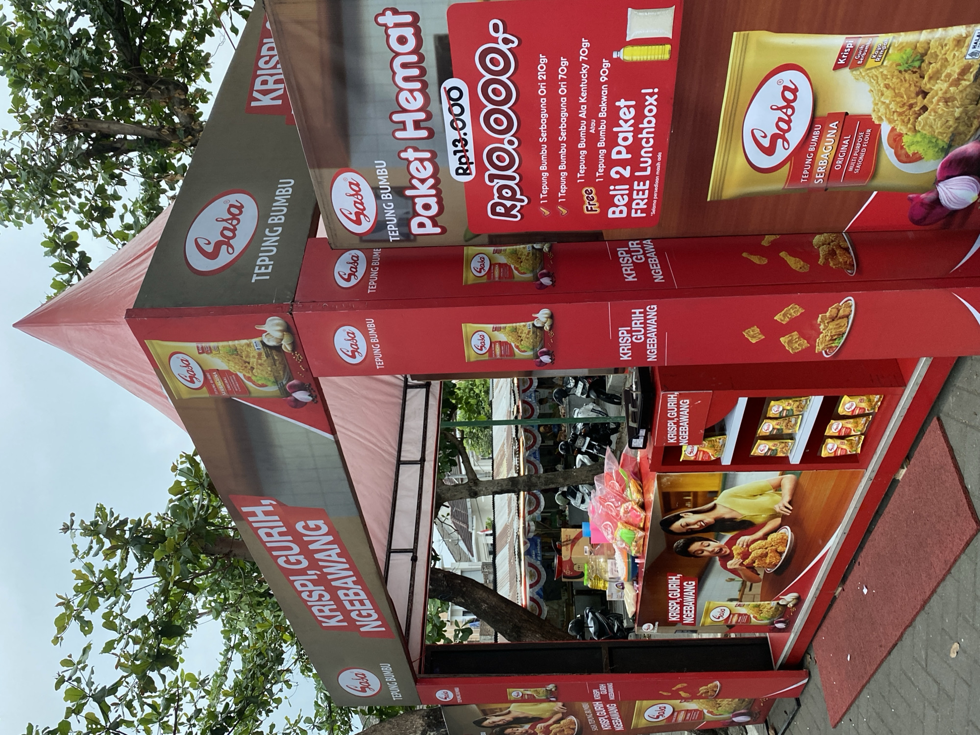 The image shows a red promotional stand for "Sasa Tepung Bumbu" (Sasa Seasoned Flour) in Ambarketawang, Indonesia. The stand is set up outdoors, with a red tent overhead and trees in the background. A sign on the stand reads "KRISPI, GURIH, NGEBAWANG," which translates to "Crispy, Savory, Aromatic." There is also a promotion offering a "Paket Hemat" (Economy Pack) for Rp10,000, which includes two packets of seasoned flour and a free lunchbox when buying two packets. Product displays, including bags of seasoned flour and images of fried chicken, are visible on the stand. Several motorcycles are parked behind the stand, and a red mat is on the ground in front of it. There are no people clearly visible in the image.