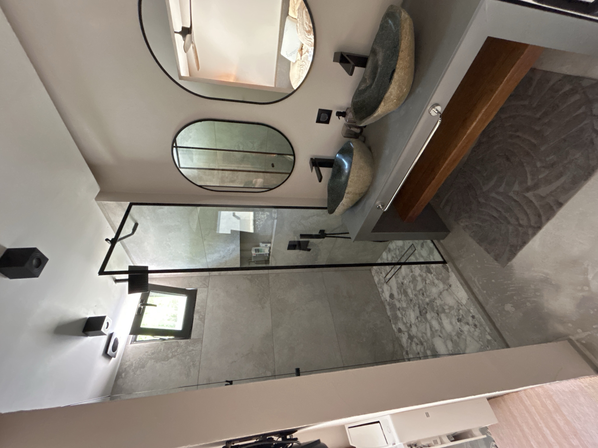 This is a photograph of a modern bathroom with a double vanity and a walk-in shower. The double vanity features two stone vessel sinks with black faucets and is set on a grey countertop. Above the vanity are two oval mirrors, one reflecting the bathroom interior and the other reflecting a bedroom scene with a bed and a ceiling fan. To the left of the vanity is a walk-in shower with grey tile walls and a marble-patterned floor. The shower is enclosed by a black metal frame and glass panels. The bathroom walls are painted a light pink color. The floor is a polished concrete. In the upper left of the image, there is a window with greenery visible outside. The scene is well-lit with recessed ceiling lights. This bathroom is located in a modern apartment in Sozopol, Bulgaria.