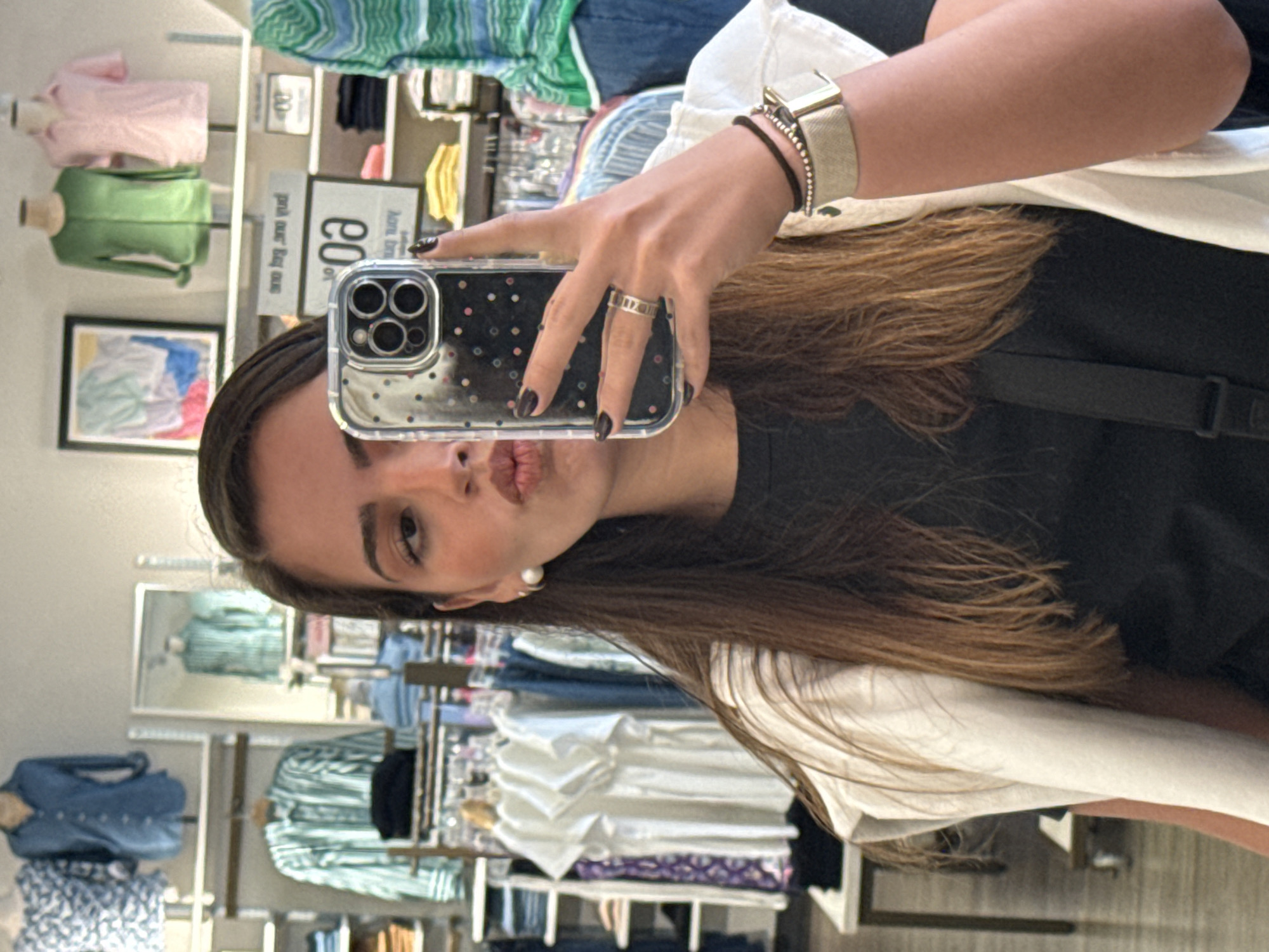 A person is taking a selfie in a retail clothing store. The individual is holding a cell phone with a clear case covered in small silver dots. The person has long brown hair, a black top, a white outer shirt, and a wristwatch. The person is puckering their lips towards the camera. Racks of folded and hanging clothing are visible in the background. The location is in San Marcos, United States of America.