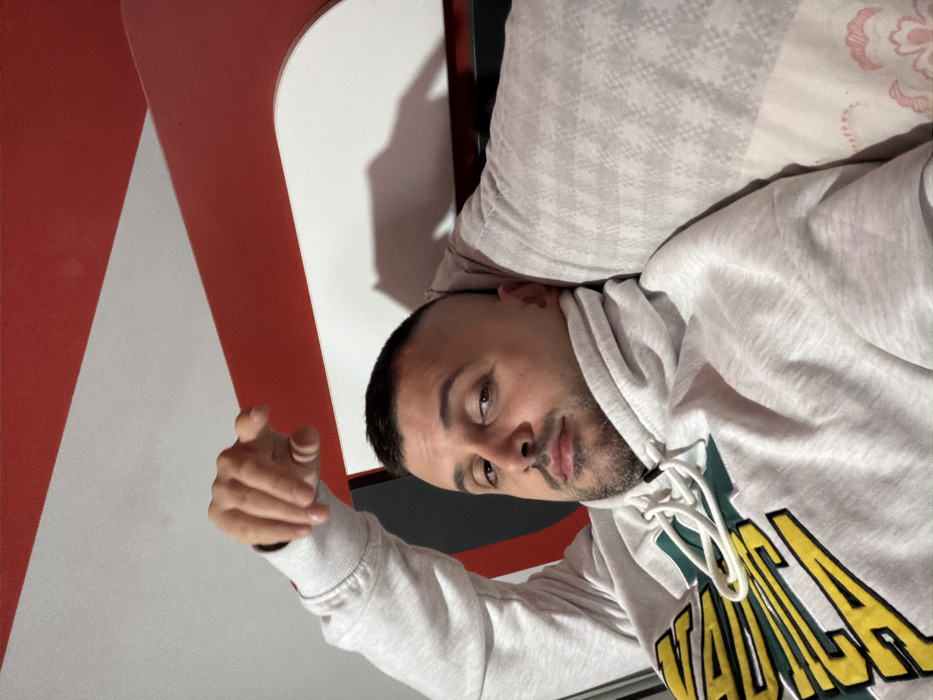 An adult male, estimated 25-45 years old, with short, faded hair and a trimmed beard, is depicted reclining indoors in Opanets, Bulgaria. He is wearing a light grey hooded sweatshirt featuring green and yellow text appliqué on the chest. His left arm is bent, with the hand positioned above his head, fingers slightly curled. His head rests against a patterned grey pillow, with a small section of floral-patterned bedding visible to the lower right. The background consists of a distinct red and white striped surface, likely a wall or headboard, exhibiting a dark shadow on the white section behind the subject's head. The subject maintains a neutral expression while looking directly at the camera.