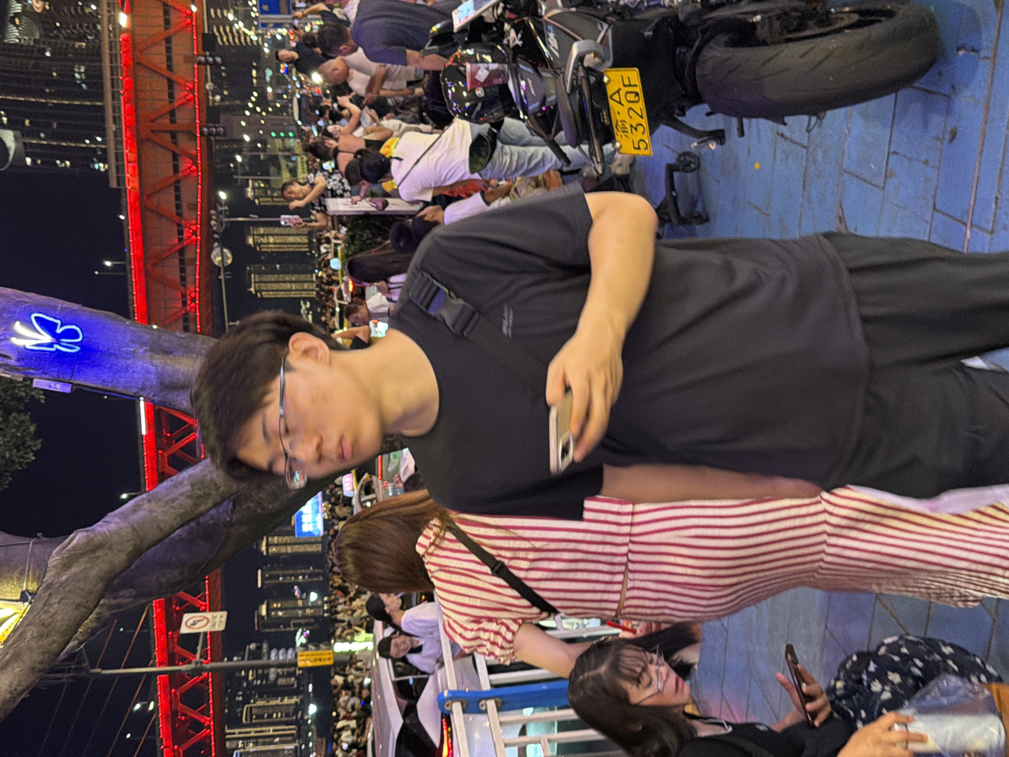 In Yuzhong District, China, a nighttime outdoor scene captures a crowd of people near a brightly lit red bridge. A young man wearing glasses and a black t-shirt holds a mobile phone in his right hand, looking to his right. To his left, a woman in a red and white striped dress stands, and a woman with glasses is seated, also holding a phone. A motorcycle with a yellow license plate that reads "渝 A 532QF" is parked on the right side of the image. The environment includes trees, streetlights, and buildings in the background, illuminated by urban lighting.