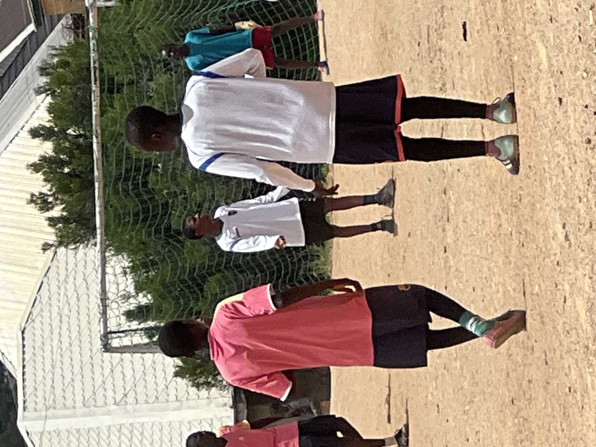The image shows a group of young people on a dirt field, likely a soccer field, in Jos, Nigeria. The foreground features two boys in athletic wear. One is wearing a pink t-shirt and dark shorts over leggings, with one foot crossed over the other, displaying a pink shoe and striped socks. The other boy is wearing a white long-sleeved shirt with blue trim and dark shorts with red trim, along with light blue athletic shoes. Behind them, two more boys are visible. One is wearing a blue t-shirt and red shorts, and another is in a white long-sleeved shirt and dark shorts. In the background, there's a soccer goal with a green net and trees, as well as a building with a white, corrugated roof. The lighting suggests it is daytime, possibly a sunny day, given the shadows on the ground.