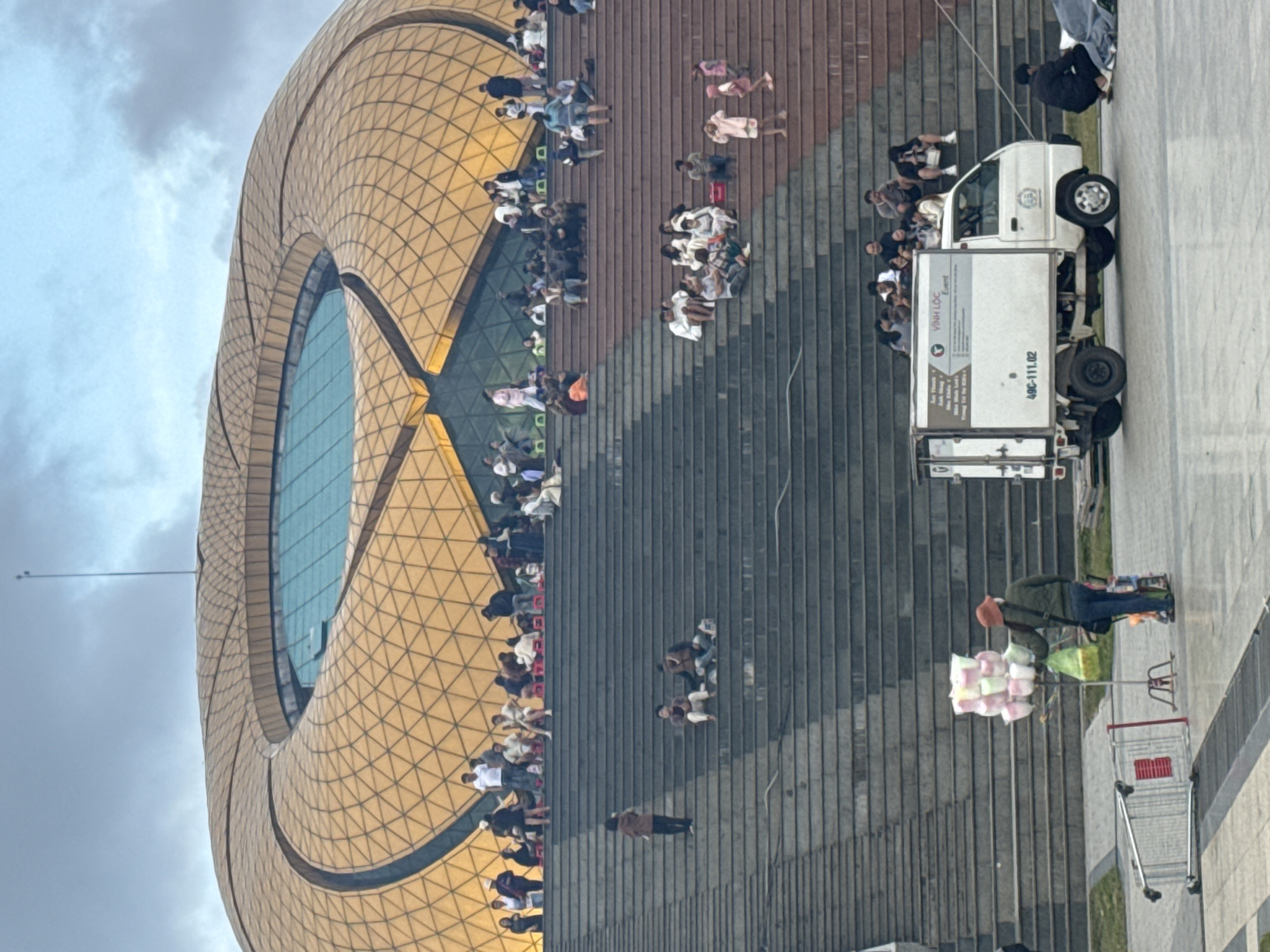 The image depicts a large, golden, geodesic-domed building with a circular opening at the top, likely a performance venue or exhibition center, situated in Da Lat, Vietnam. A wide, tiered staircase leads up to the building, populated by numerous people of various ages who are seated, standing, and interacting. The crowd is diverse, with individuals dressed casually, some appearing to be tourists or locals enjoying the open space. In the foreground, a street vendor is visible with a cart holding several colorful cotton candy puffs, while a white delivery truck with Vietnamese license plates is parked nearby. The overall atmosphere suggests a public gathering or event. The sky is overcast with scattered clouds.