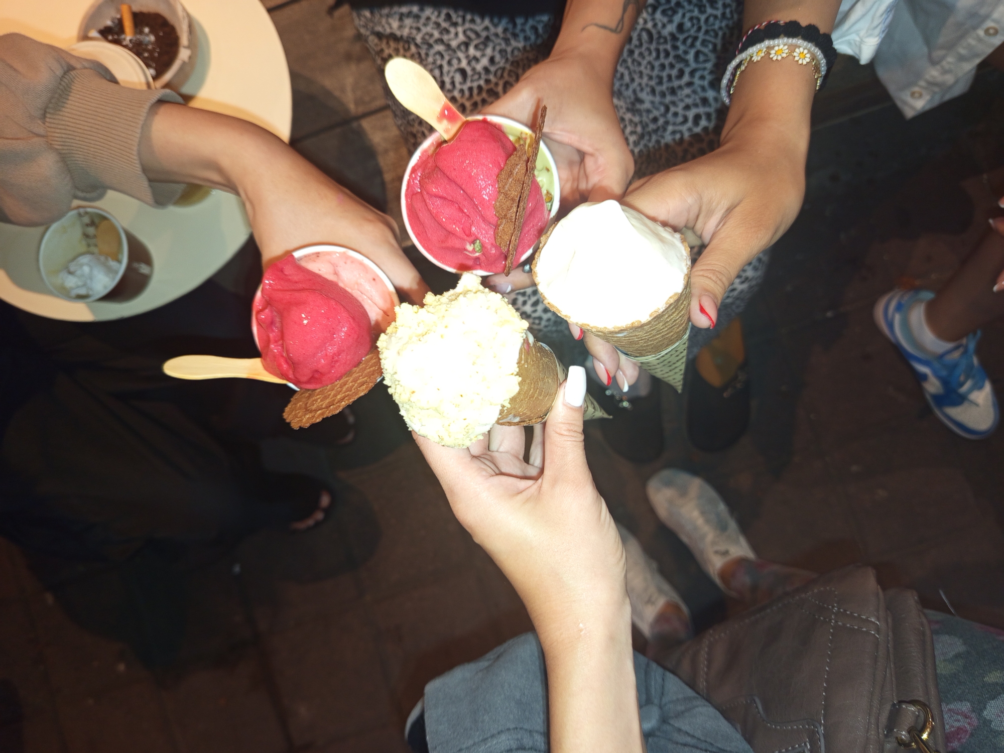 A group of people is standing in what appears to be an outdoor setting in North Kuta, Indonesia, holding ice cream. There are five individuals visible, each holding either a cup or cone of ice cream. The ice cream flavors include red, white, and yellow. Some of the ice cream cups have wooden spoons. One person is wearing a daisy bracelet and has red nail polish on their index finger. Another person wears blue and white sneakers. A brown leather bag is visible. There is a small round table with two cups. One cup has a cigarette.