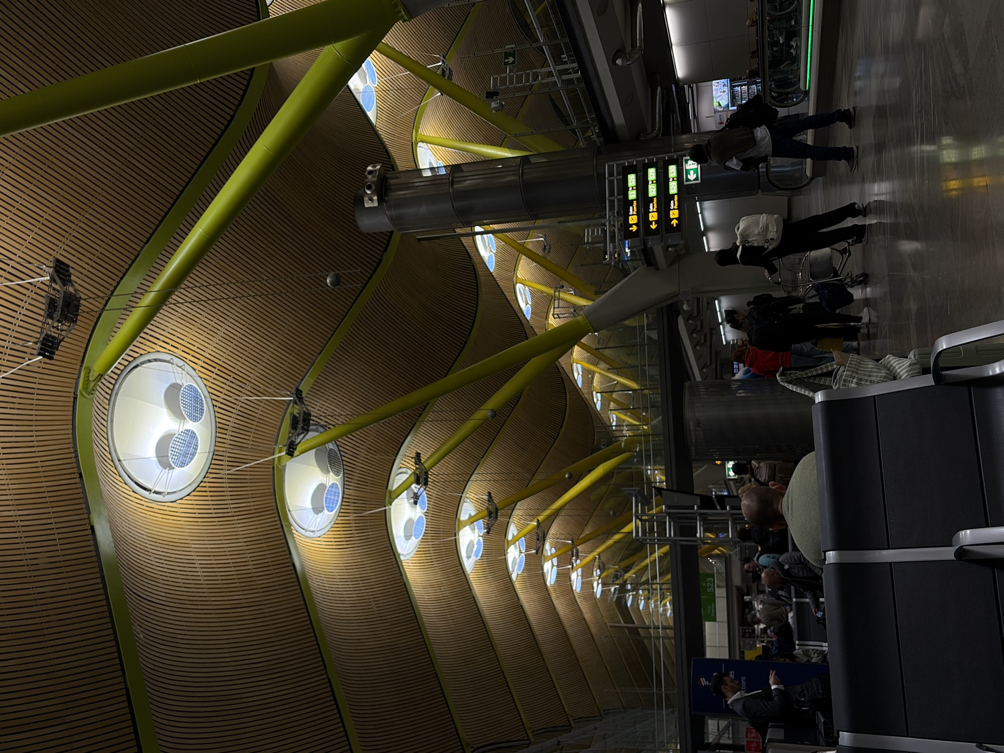 The image captures the interior of Madrid-Barajas Airport, specifically Terminal 4. The architecture is notable, featuring a curved wooden ceiling supported by green beams, punctuated by circular skylights. People are present, some with luggage carts, moving through the terminal. The setting appears to be a waiting area with seating visible on the right side of the image. Overhead, directional signs guide passengers.