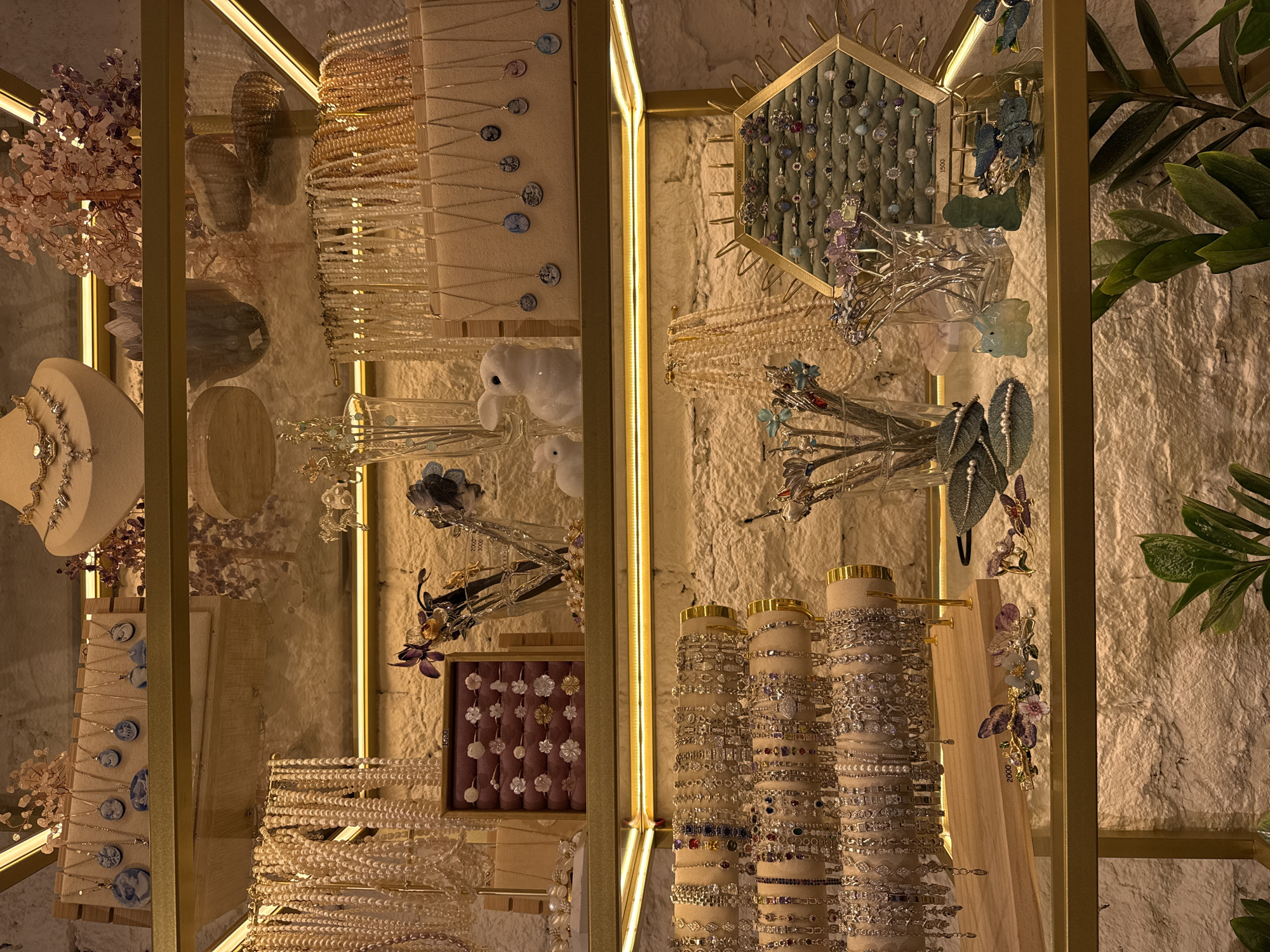 The image shows a display of jewelry in a store, likely in Saint Petersburg, Russia. The store features several shelves filled with various accessories. The top shelf displays necklaces and pendants, some with blue cameo-like designs. Below that, pearl necklaces and more pendants are showcased. The third shelf is dedicated to bracelets, arranged on tiered stands. The bottom shelf features hair accessories, rings displayed on a hexagonal tray, and small decorative figurines. The overall aesthetic is elegant, with warm lighting highlighting the merchandise.