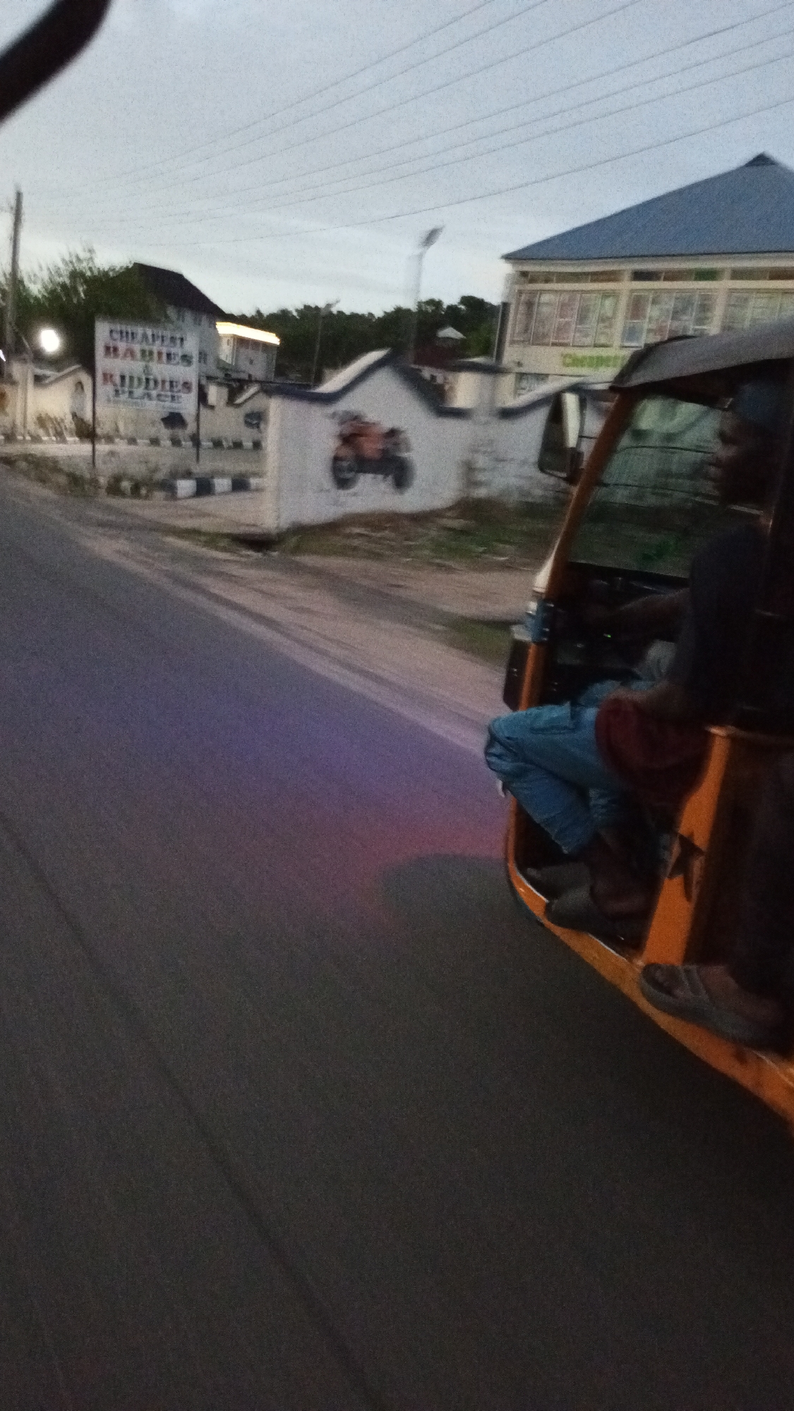 The image, likely taken from a moving vehicle, shows a scene on a street in Lokoja, Nigeria. A man is driving a three-wheeled yellow passenger vehicle, commonly known as a "keke" or "tricycle taxi". The focus is on the right side of the road, where buildings and walls are visible. One building has a sign that reads "CHEAPEST BABIES & KIDDIES PLACE". A motorcycle mural is painted on a wall. The sky is overcast. The road appears to be paved and is marked with a purple streak.