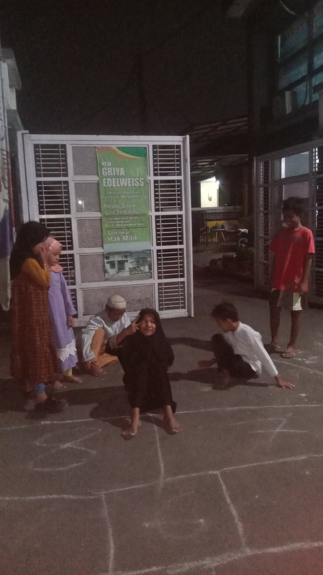 In Sumberjaya, Indonesia, a group of four children and one adult are gathered on a paved outdoor surface. The surface is marked with chalk lines, suggesting a game is in progress. The lighting is dim, indicating it is likely nighttime or a very overcast day.
A young girl in the foreground, wearing a dark headscarf and dress, is sitting and making a playful gesture with her fingers near her eye. To her left, an adult male wearing a white cap and traditional clothing is also seated. Two young girls are standing to the left of the seated individuals. One is wearing a patterned orange and brown dress, and the other is in a purple dress and pink headscarf. Further to the right, a young boy in a white long-sleeved shirt and dark pants is crouching on the ground, appearing to be part of the game. In the background, a teenage boy in a red t-shirt and shorts stands facing left.
Behind the group, a large white structure resembling an air conditioner unit or decorative screen is visible. A green sign with white text is attached to it, advertising "NEW GRIYA EDELWEISS" with additional details about housing, investment, and property ownership. The environment appears to be an urban or semi-urban area, with glimpses of buildings and what might be a fence or gate in the background. The overall scene captures a moment of outdoor activity among children in a residential setting.