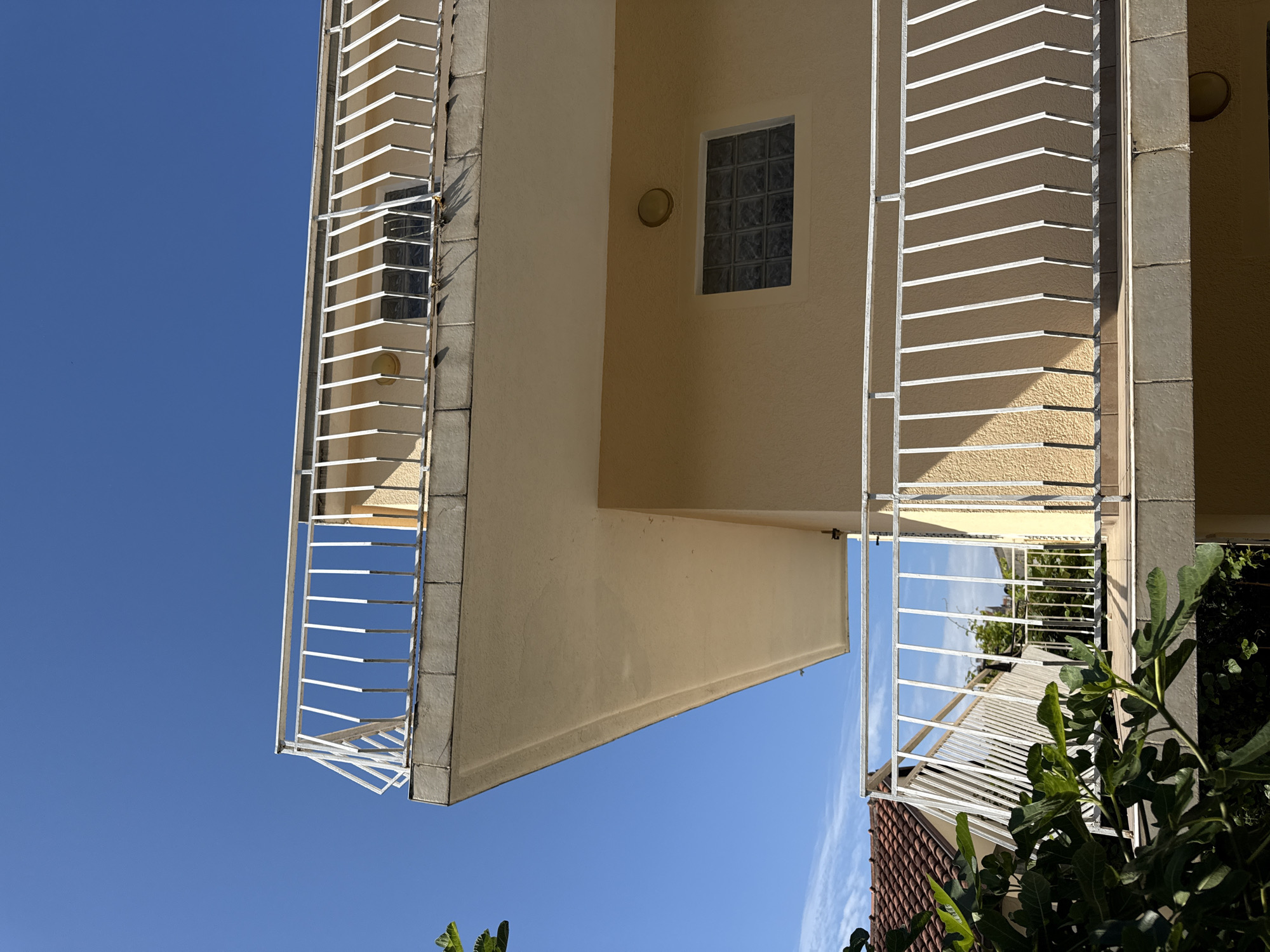 An outdoor, sunlit architectural photograph shows a cream-colored building, likely a residential complex or hotel, set against a bright, clear blue sky. The image focuses on the building's exterior, featuring multiple levels of balconies with distinctive white metal railings. These railings consist of a series of staggered horizontal slats, casting prominent shadows on the textured facade of the building, indicating strong sunlight.
One section of the building prominently displays a window composed of frosted glass blocks, with a circular wall light positioned nearby. The building's walls are finished in a light cream or sandy yellow stucco. To the right, a darker, possibly gray-tiled or stone-clad column separates parts of the structure.
At the lower right edge of the frame, lush green foliage, possibly from a fig tree, is visible, partially obscuring a terracotta-tiled roof from a lower structure or an adjacent part of the same building. The sky is largely cloudless, with only faint wisps of white visible in some areas. No people or visible text are present in the image. The scene suggests a warm, sunny day in a location like Budva, Montenegro, consistent with the architectural style and vegetation.
