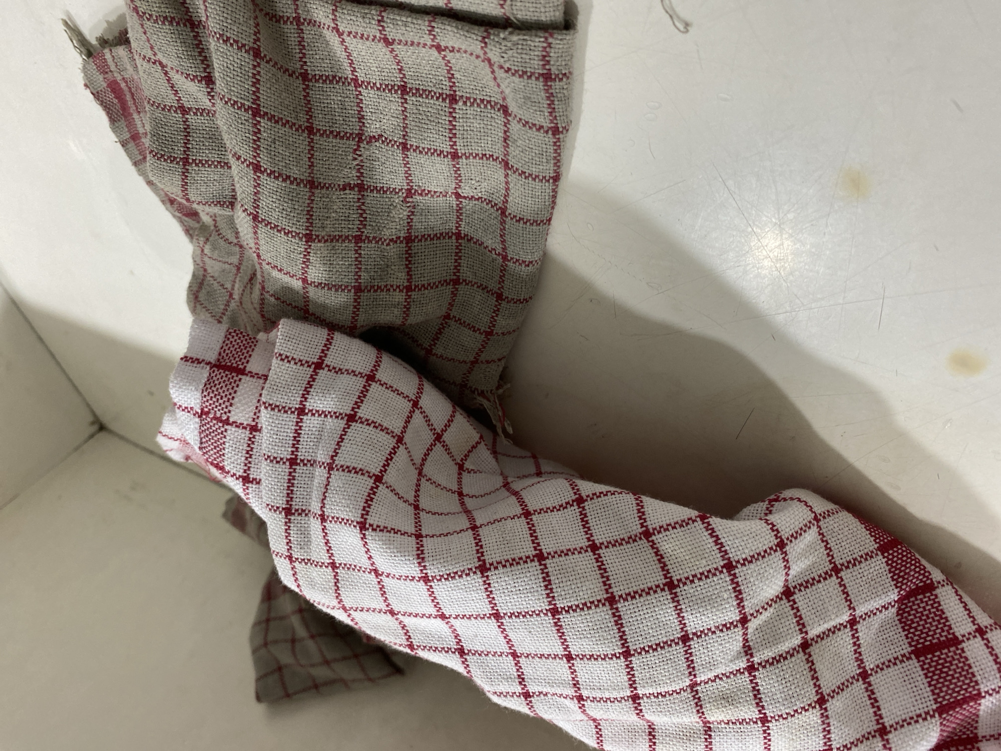 Two dishtowels, one white with red plaid and the other gray with red plaid, are resting on a white surface. The towels are bunched up and appear to be made of a textured fabric. The white towel is positioned to the left and is partially obscuring the gray towel, which is to the right. The background is a neutral, light-colored surface, possibly a countertop or shelf. The lighting is dim, casting subtle shadows. There is a slight yellow stain on the white surface in the lower right quadrant of the image. The scene is indoors and appears to be in a domestic setting.