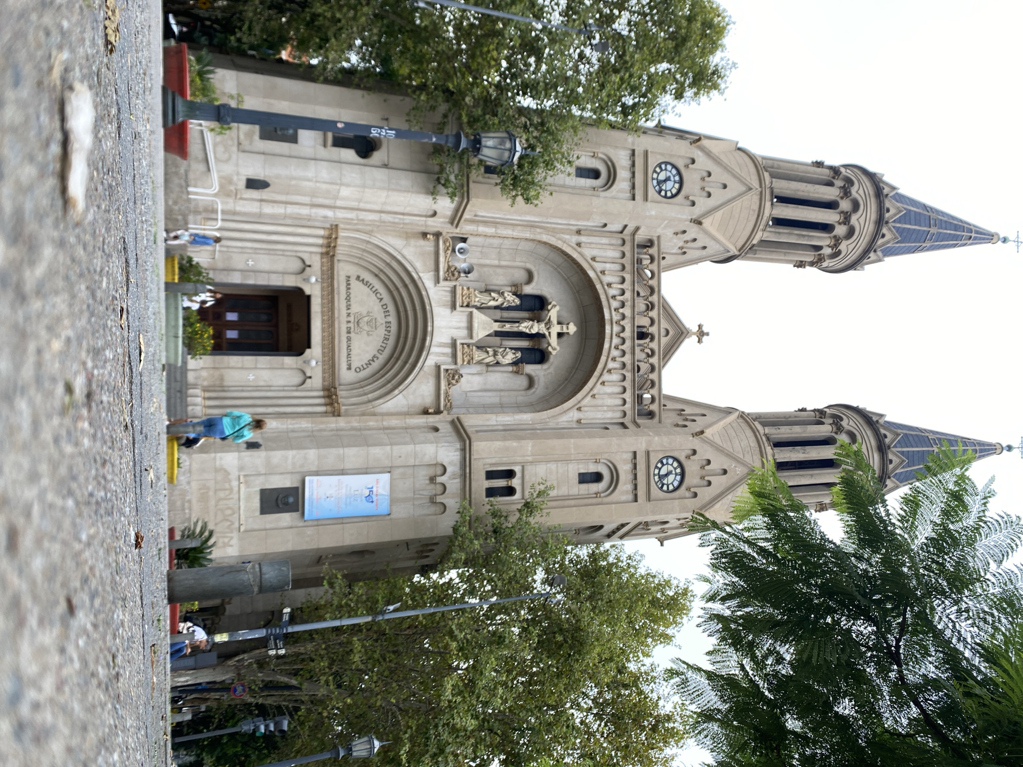 The image presents the Basílica del Espíritu Santo, also known as Parroquia Nuestra Señora de Guadalupe, situated on a street in Buenos Aires, Argentina. The church's facade, constructed from light-colored stone, features two prominent, symmetrical bell towers, each topped with a dark, pointed spire and a cross. A circular clock face is visible on each tower. The central entrance consists of a large arched portal, above which a sculpture of the Crucifixion is flanked by two additional statues. Inscriptions identify the structure as "BASILICA DEL ESPIRITU SANTO" and "PARROQUIA N. S. DE GUADALUPE." A blue promotional banner and a bronze plaque are affixed to the right side of the facade.
The scene is captured from a low vantage point, with a paved ground surface and scattered debris visible in the immediate foreground. Lush green trees partially obscure parts of the church, with a prominent branch featuring fern-like leaves extending into the upper right portion of the frame. Several individuals are visible near the church entrance and on the surrounding sidewalk: one person in a light blue top and jeans stands facing away from the camera on the right side of the steps; another in a white top is partially visible closer to the main entrance on the left steps; a third person in a blue shirt is positioned further left on the steps. On the right, a person in a white top is seated on a yellow planter, and another individual is visible further down the street. The environment includes black ornate lampposts, a gray utility pole with street signs and a traffic light, red and yellow planters, and metal railings. The sky is overcast and light gray, providing diffused natural light.