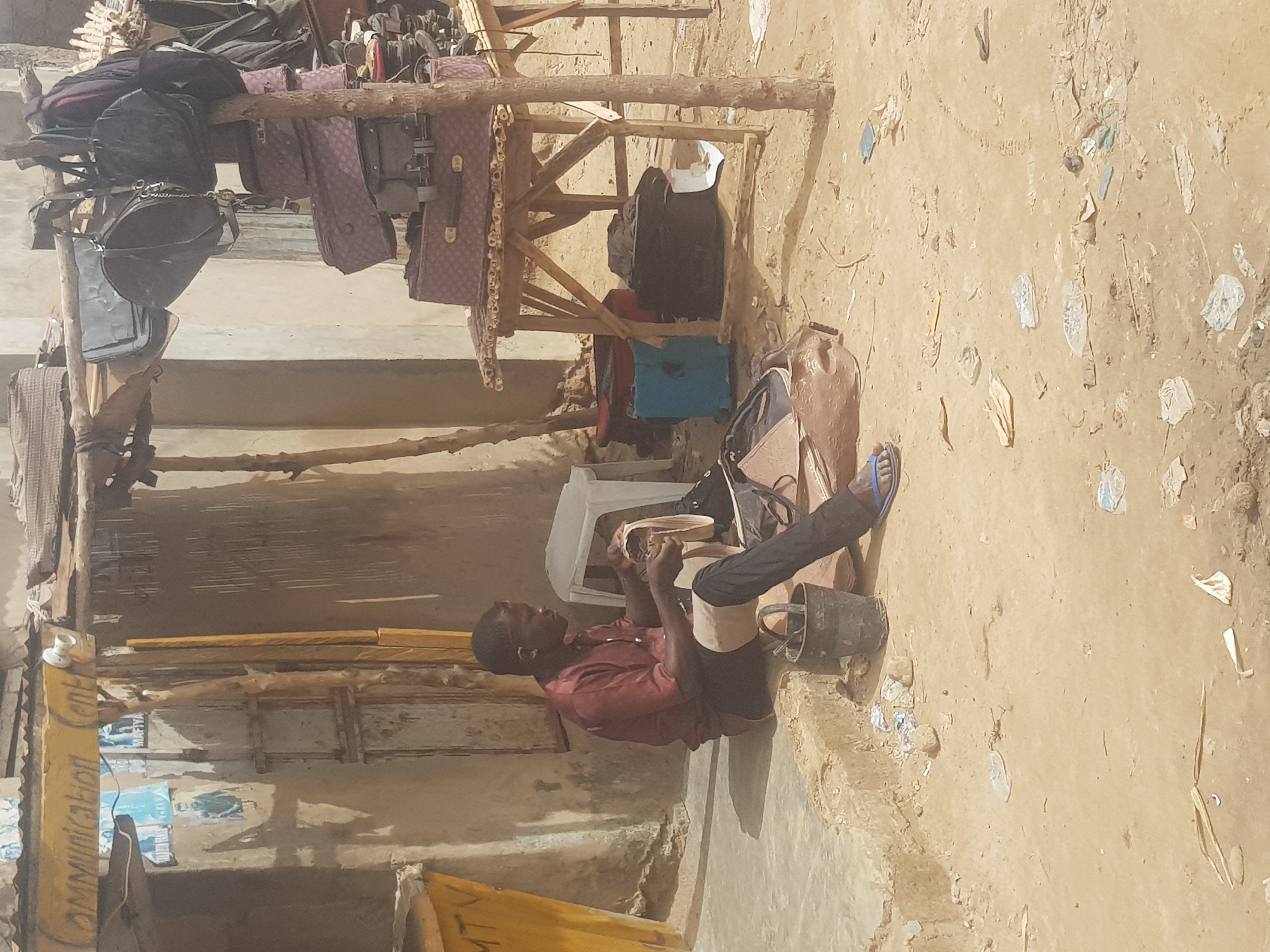 A man sits in front of a building in Dutsinma B, Nigeria. He is wearing a red shirt, dark pants, and blue flip-flops, and appears to be reading or working on something he holds in his hands. A white plastic chair supports him, and a black bucket is nearby. Above him, a wooden structure displays an assortment of luggage, bags, and cases for sale. The ground is sandy and littered with debris. The building behind him has a sign partially visible that reads "Communication Center".